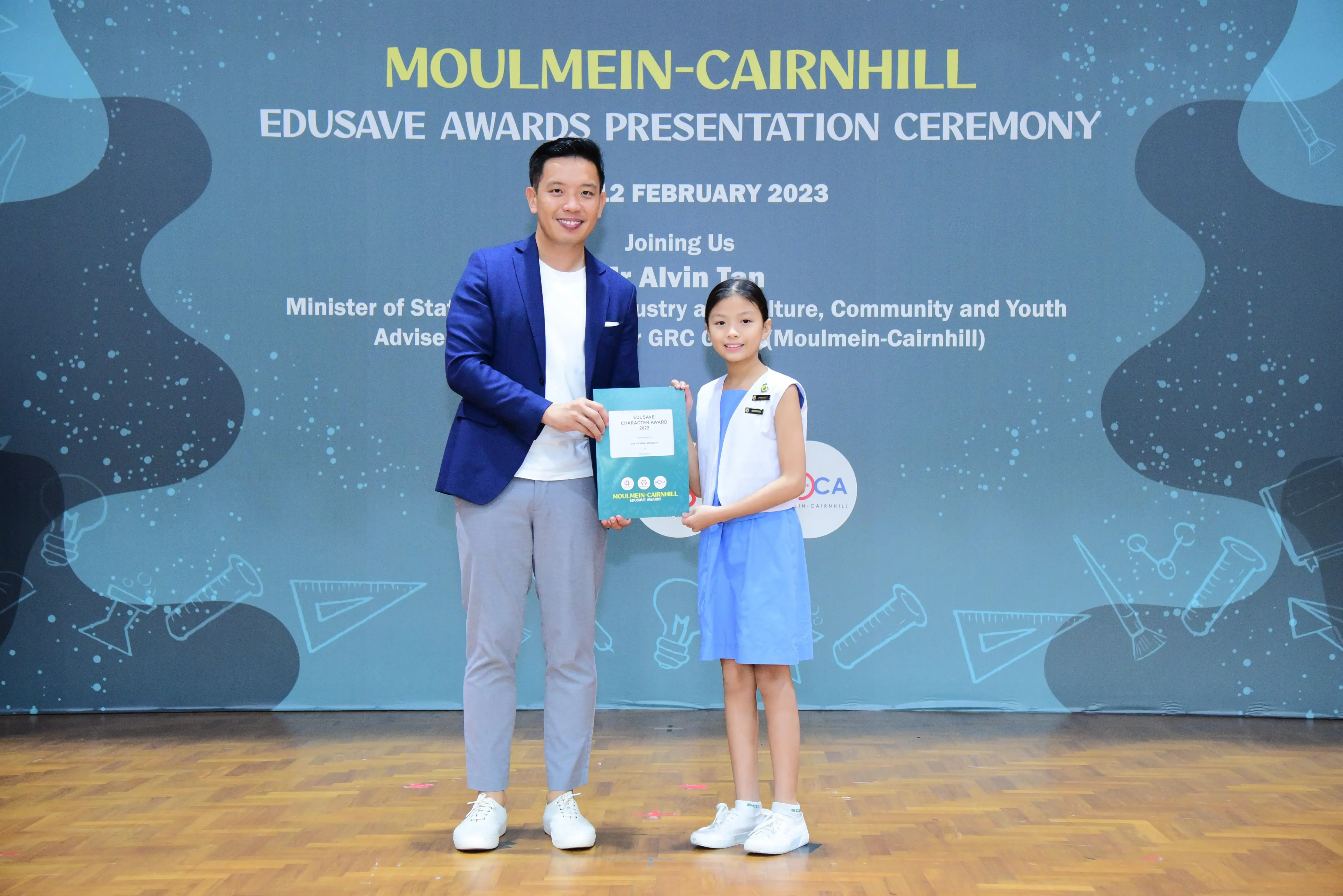 A man in a blue blazer and a young girl in uniform hold an award at the Moulmein-Cairnhill Edusave Awards Ceremony 2023.