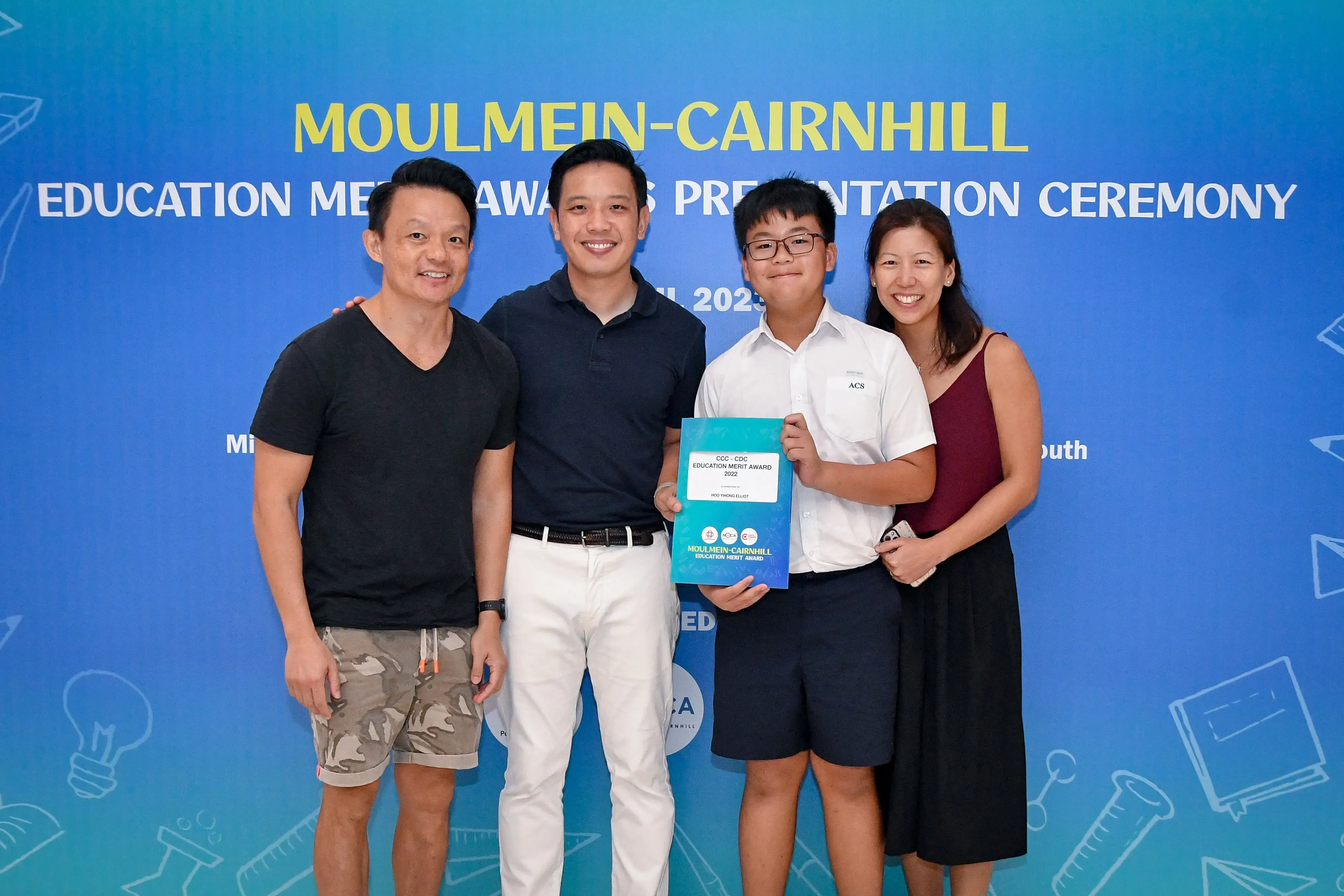 Four people smile in front of a Moulmein-Cairnhill Education Merit Awards banner; a boy holds a certificate.