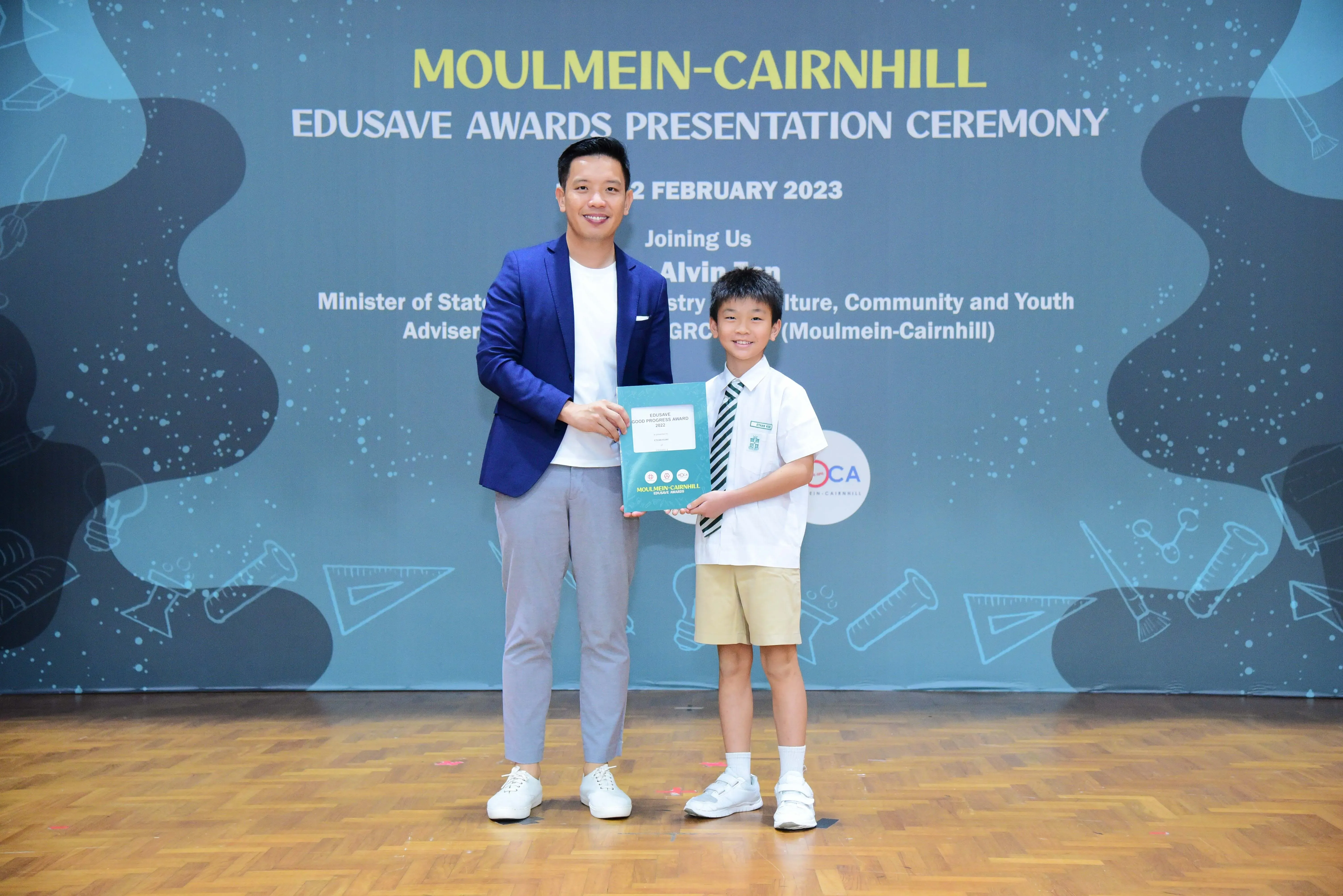 A man in a blue blazer and a young boy in school uniform hold a certificate at the Moulmein-Cairnhill Edusave Awards 2023.