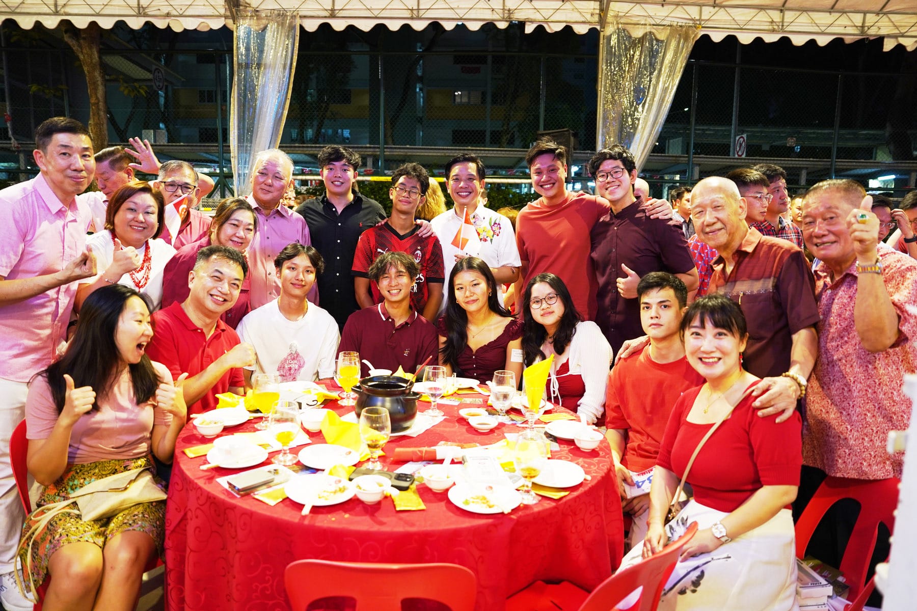 A large group of smiling people pose around a round table with a red tablecloth at an outdoor dinner event.