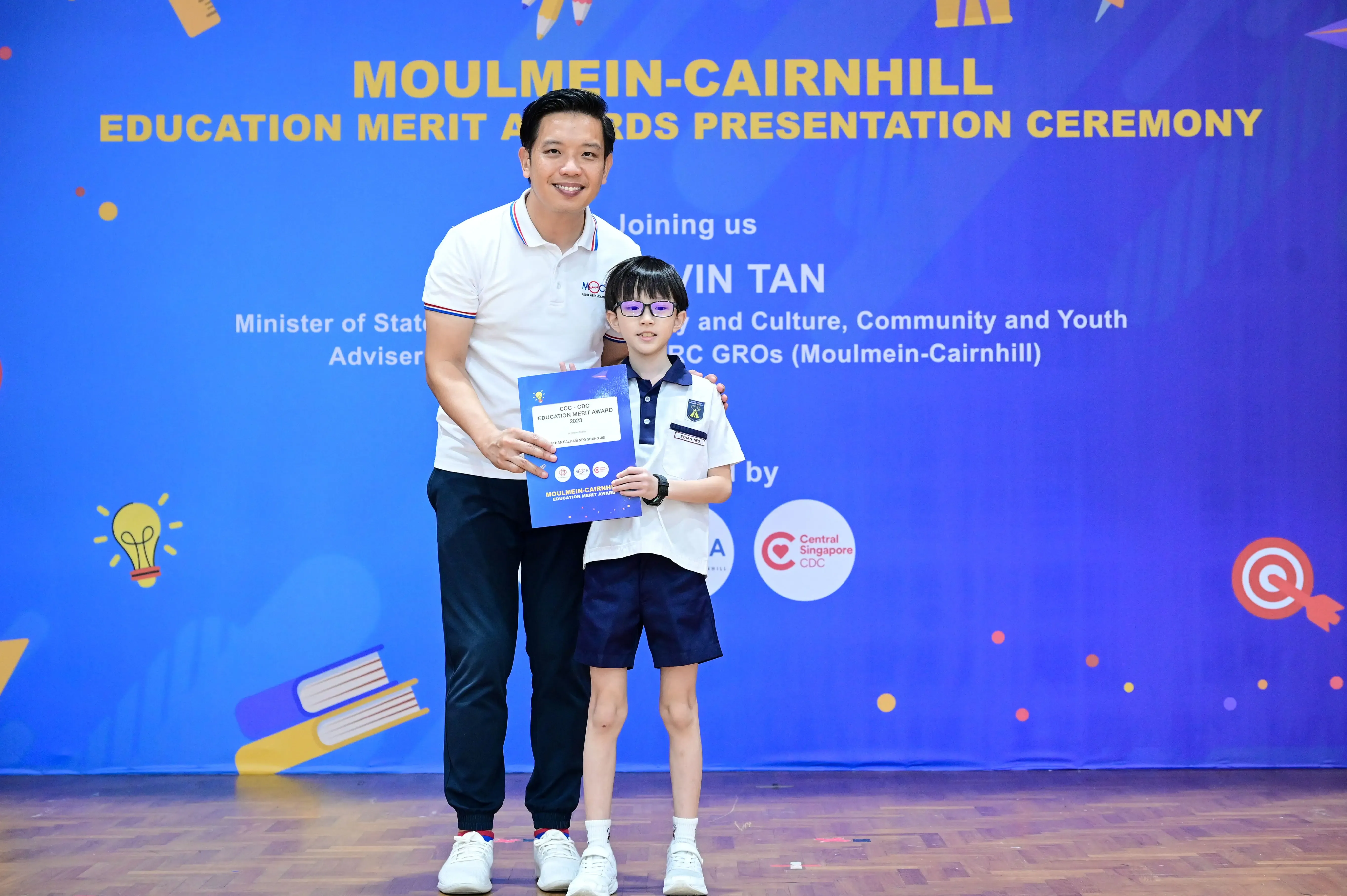A man and a young boy holding a certificate stand before a Moulmein-Cairnhill Education Merit Awards banner.
