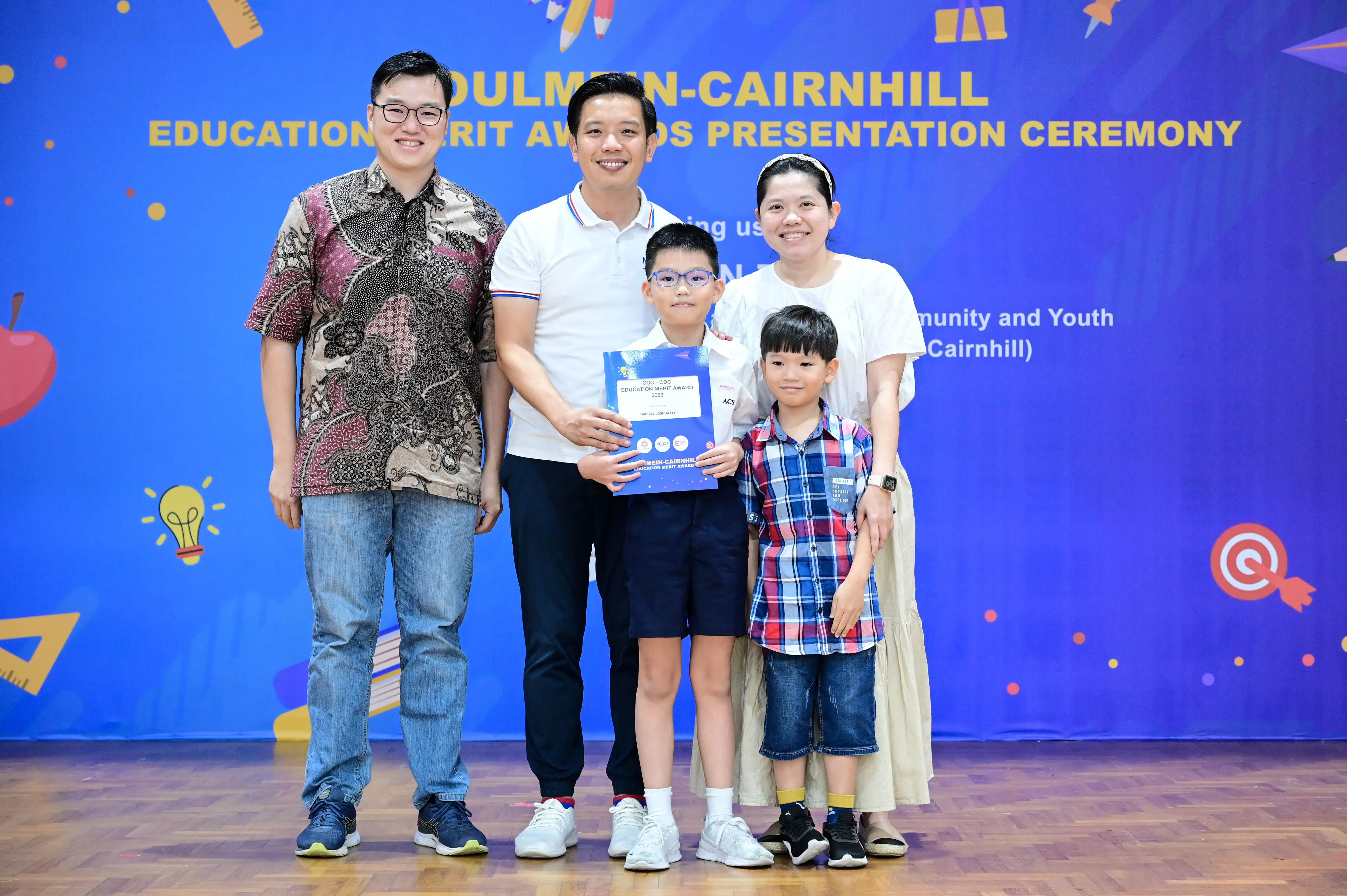 Five people pose smiling before a blue Education Merit Awards Presentation Ceremony banner, with a boy holding a certificate.