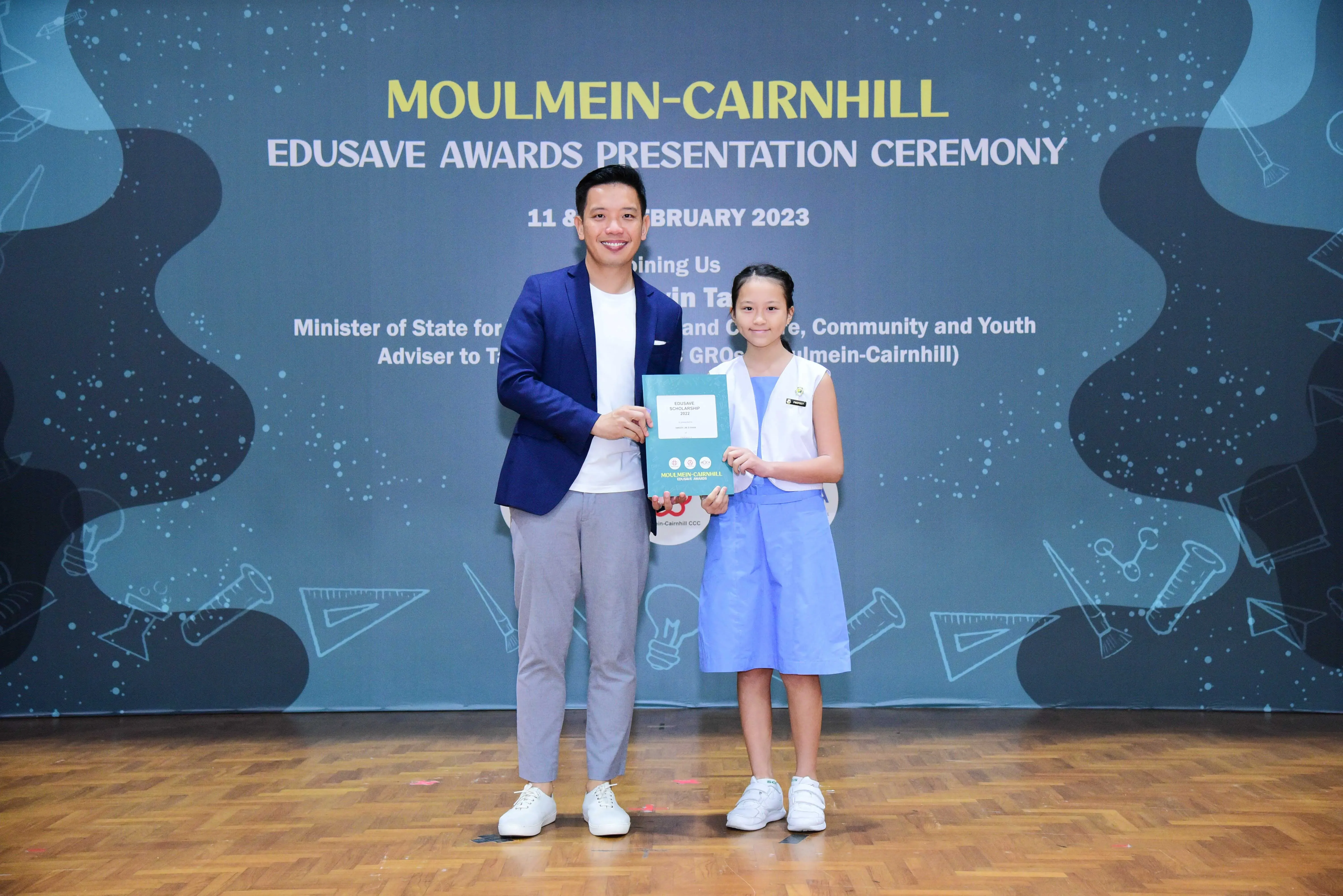 A man and a young girl hold a certificate together in front of a Moulmein-Cairnhill Edusave Awards banner.