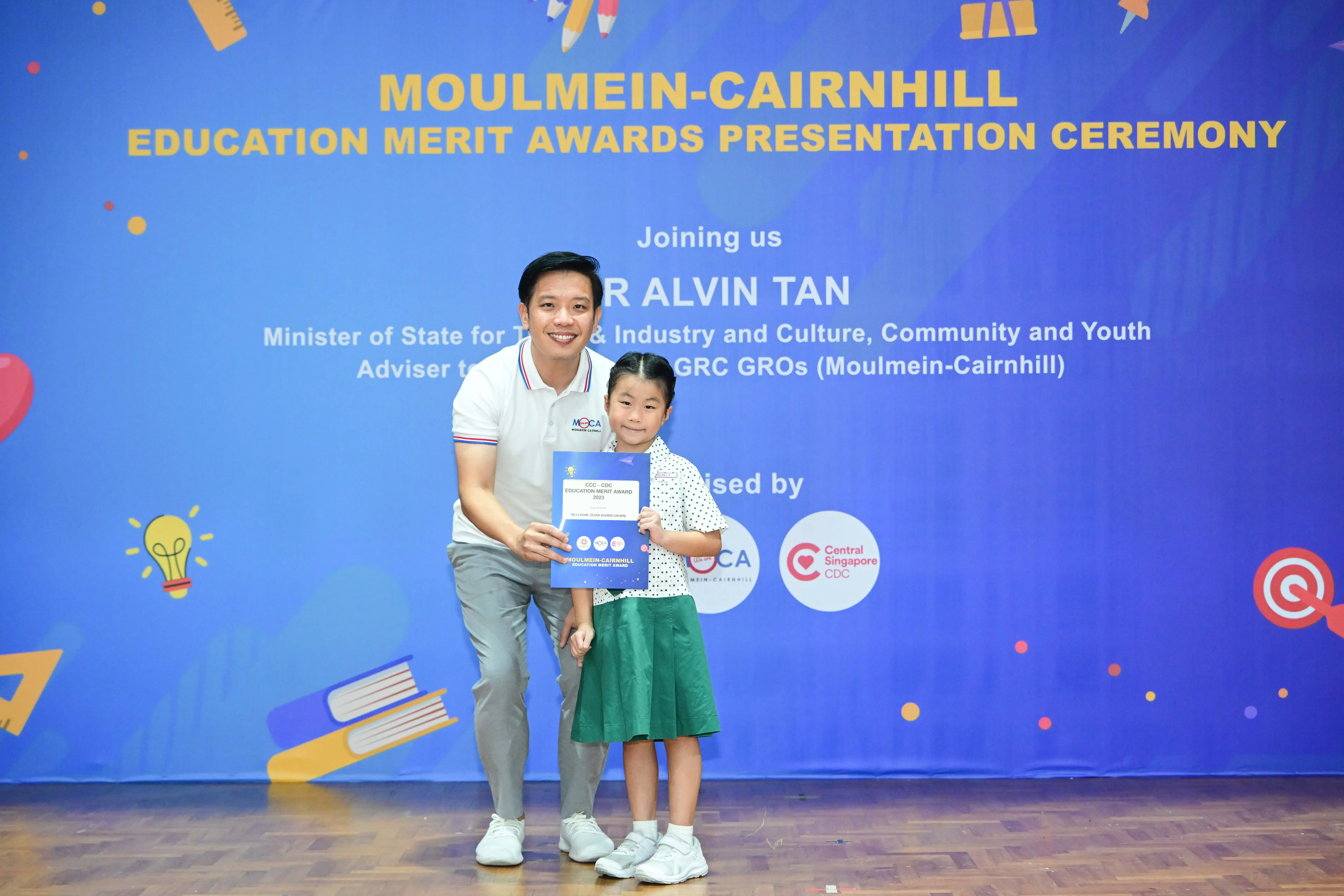 A man and a young girl pose holding a certificate in front of a Moulmein-Cairnhill Education Merit Awards banner.