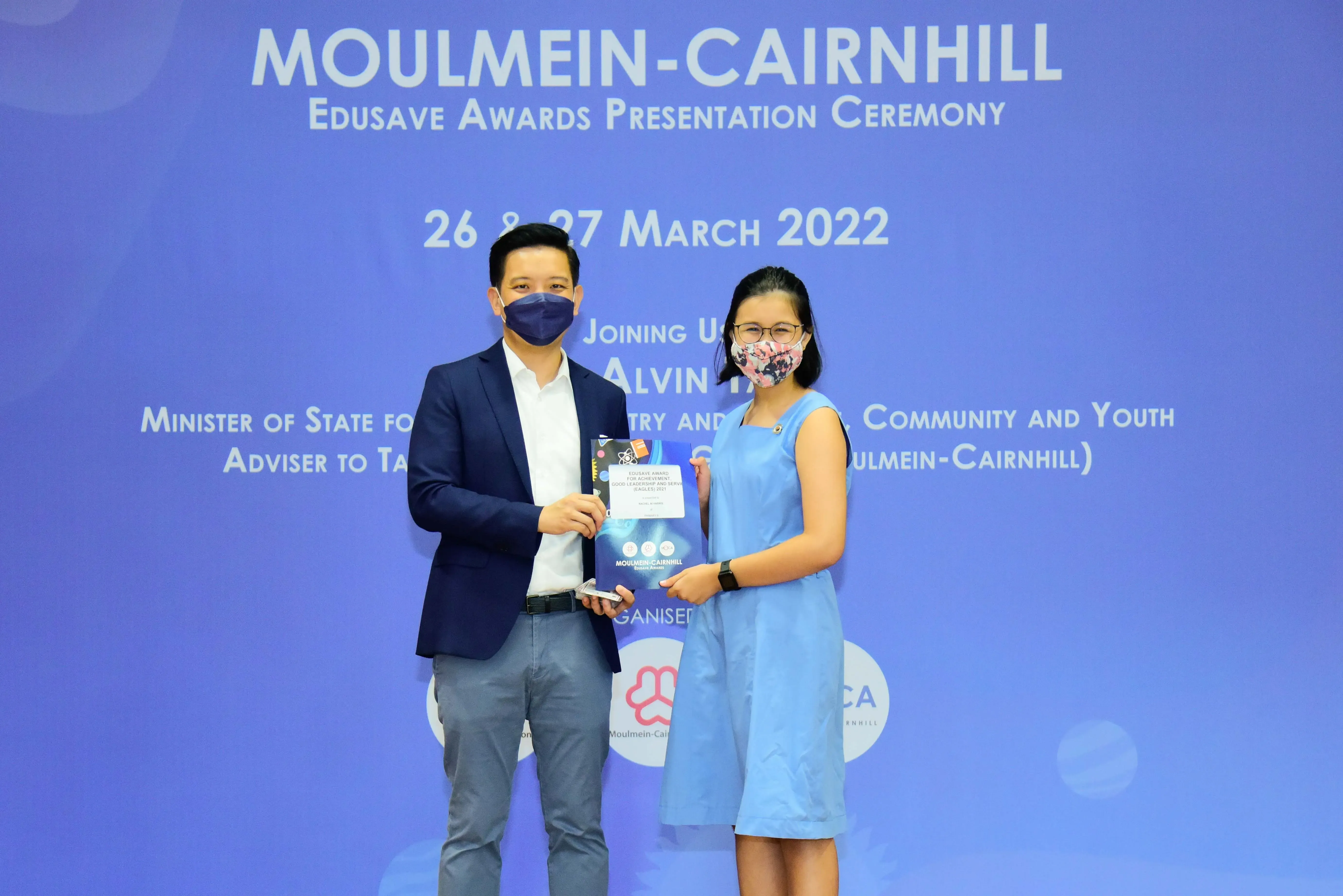 Two masked people hold an award at the Moulmein-Cairnhill Edusave Awards Presentation Ceremony, March 2022.