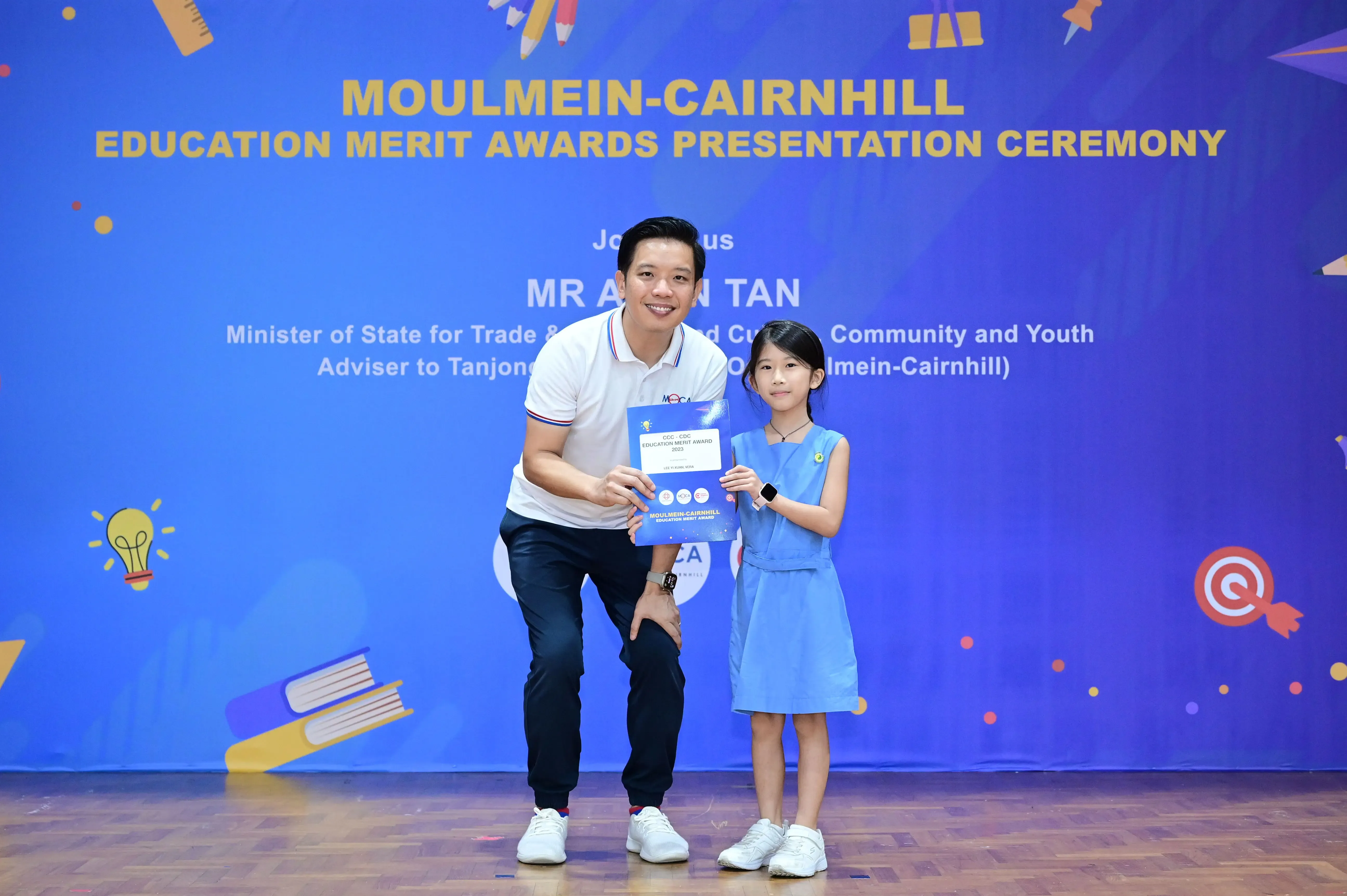 A man and a young girl hold a certificate together in front of a Moulmein-Cairnhill Education Merit Awards banner.