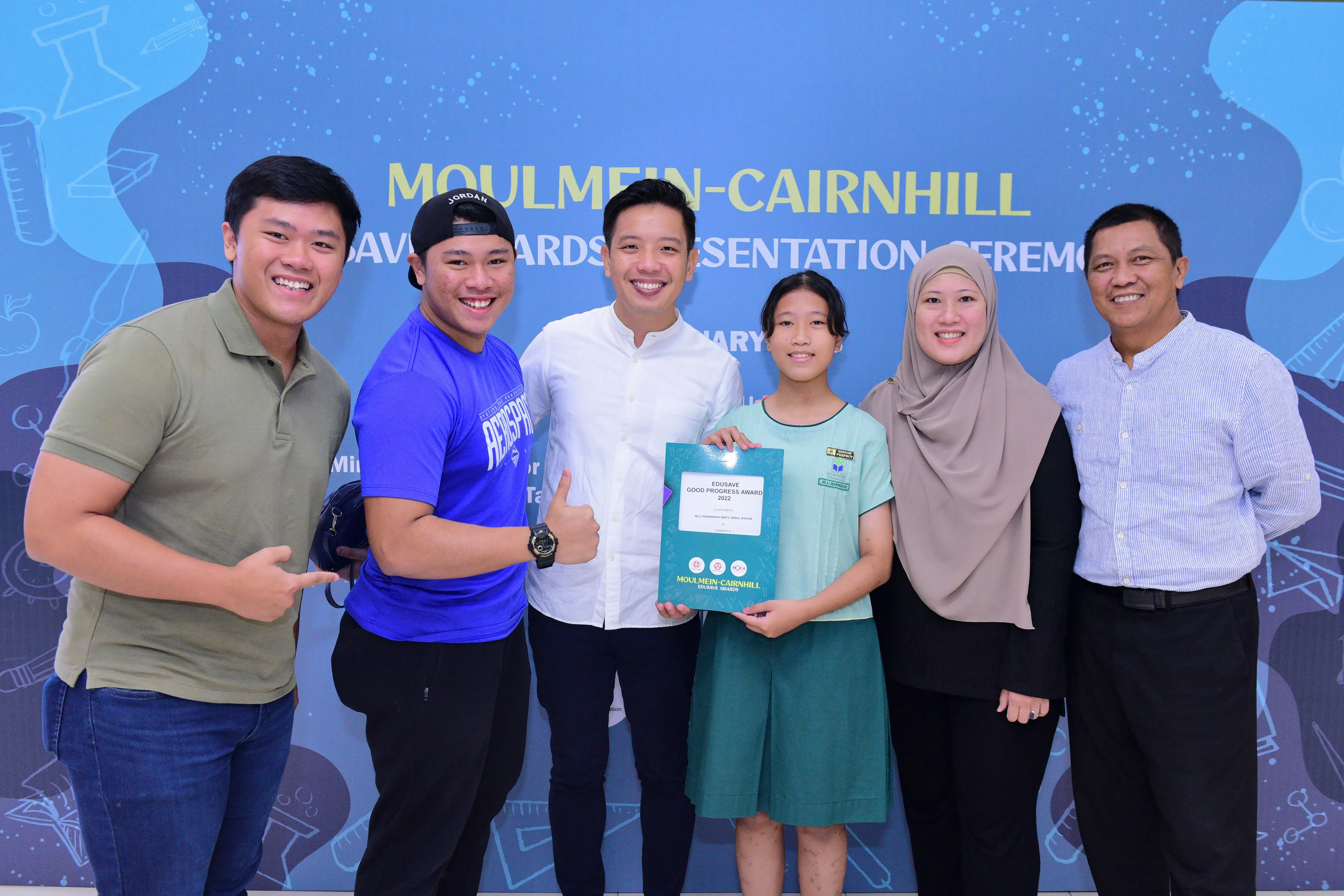 Six people smile at a Moulmein-Cairnhill Edusave Awards ceremony, with a girl holding an award certificate.