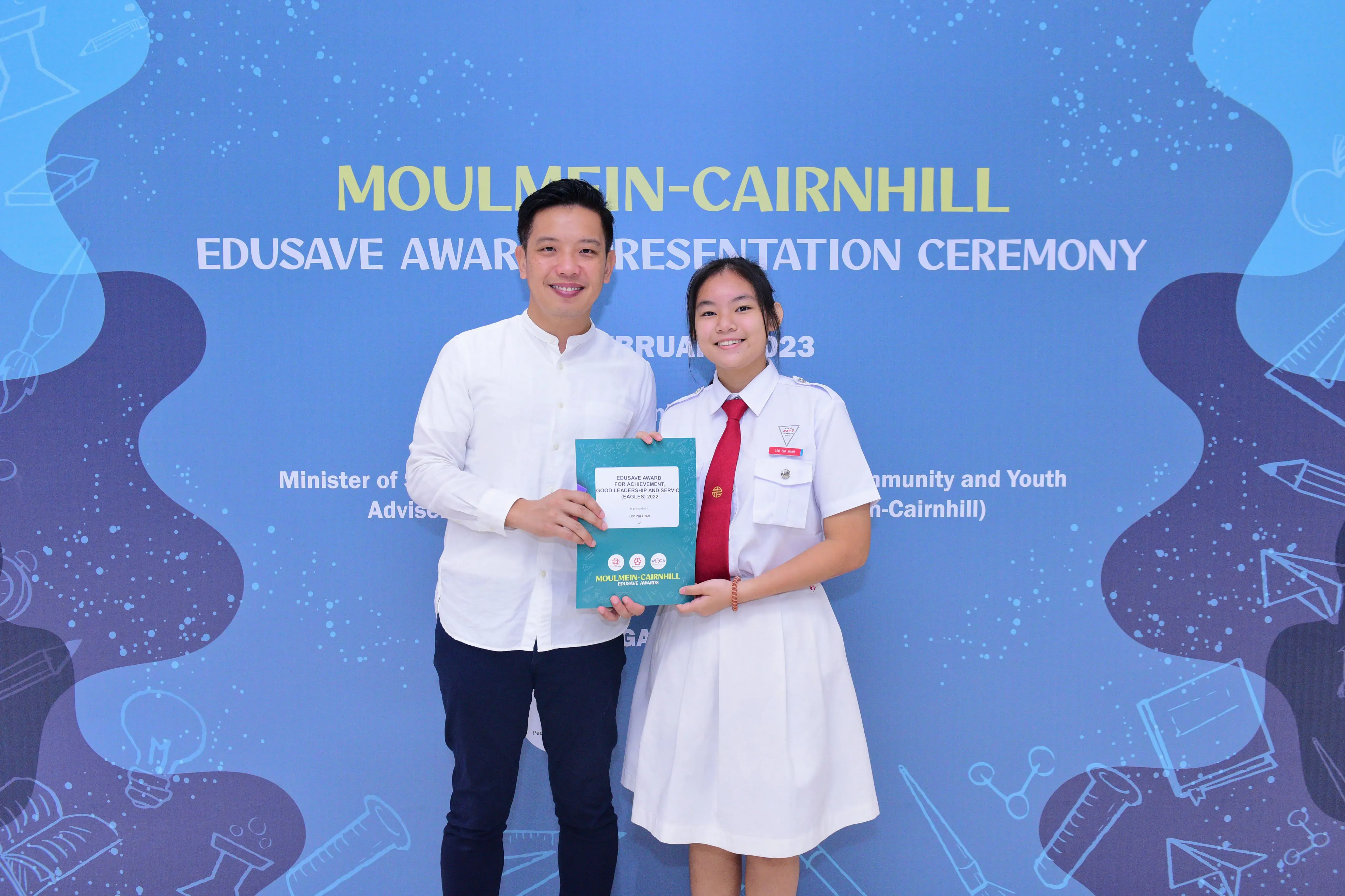 A man and a student in school uniform hold an award booklet at the Moulmein-Cairnhill Edusave Award Ceremony 2023.