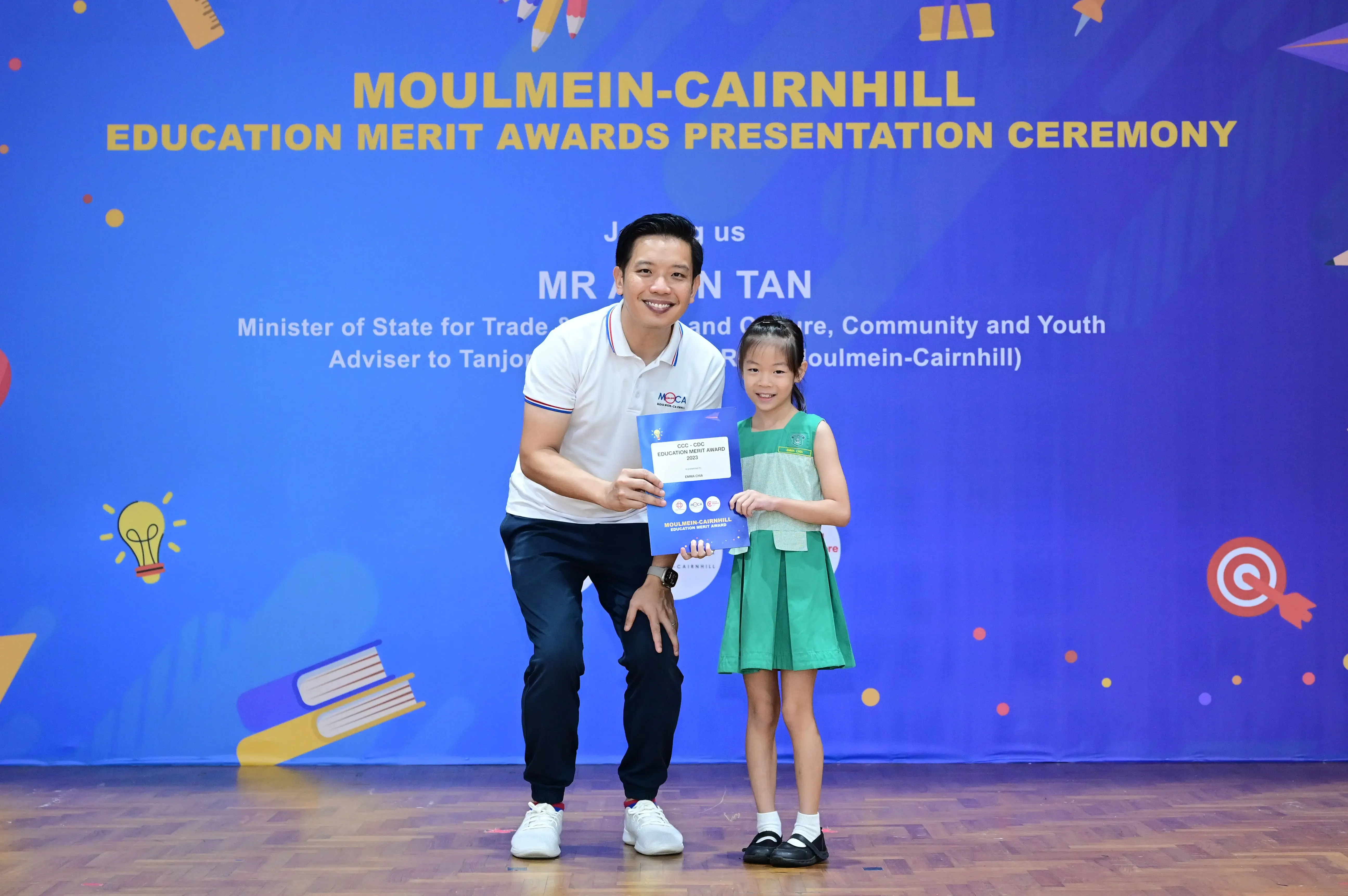 A man and a young girl hold a certificate in front of a Moulmein-Cairnhill Education Merit Awards banner.