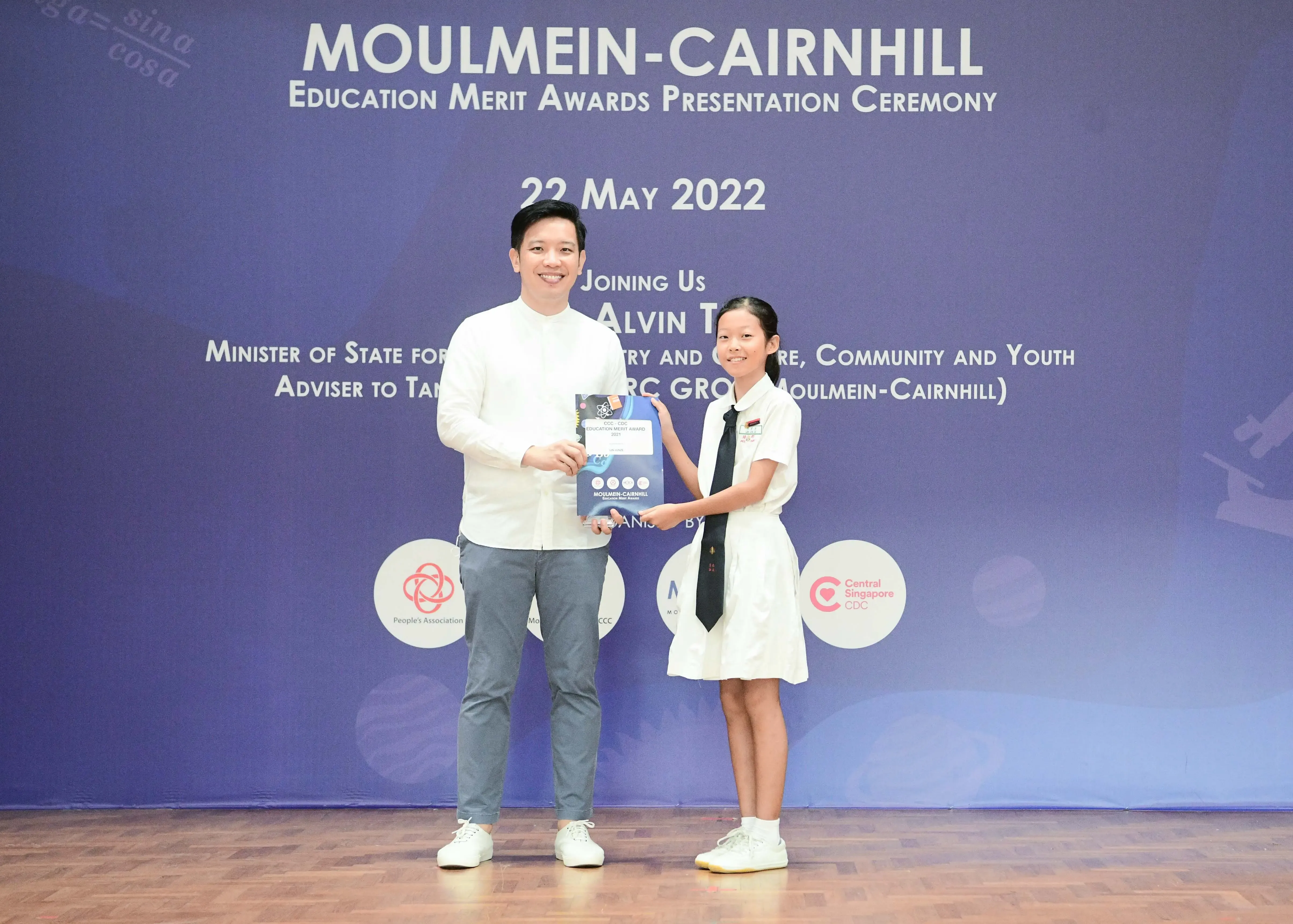 A man and a young girl in school uniform hold an award together at the Moulmein-Cairnhill Education Merit Awards ceremony.