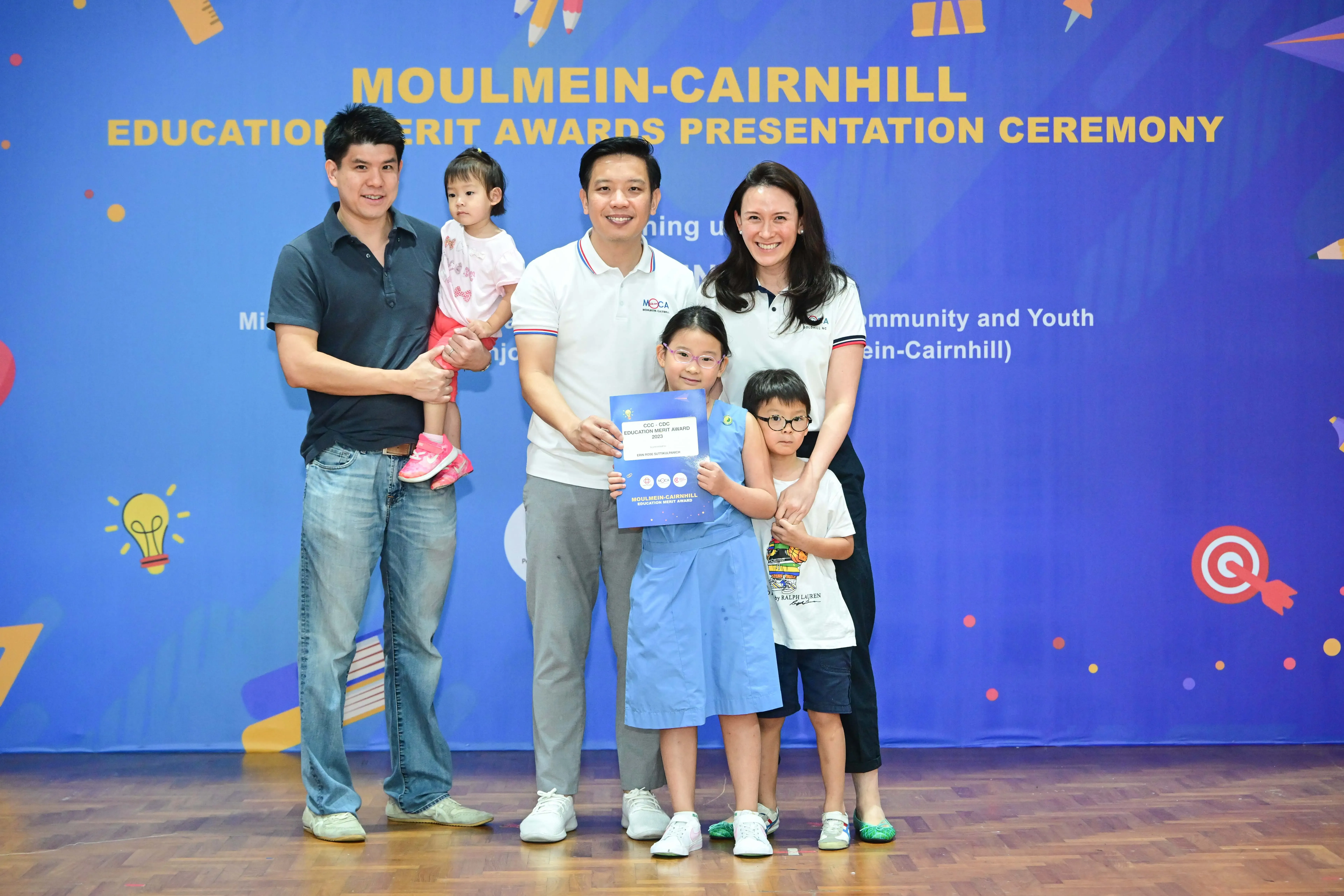 Five people pose before a Moulmein-Cairnhill Education Merit Awards banner, with a girl holding a certificate.