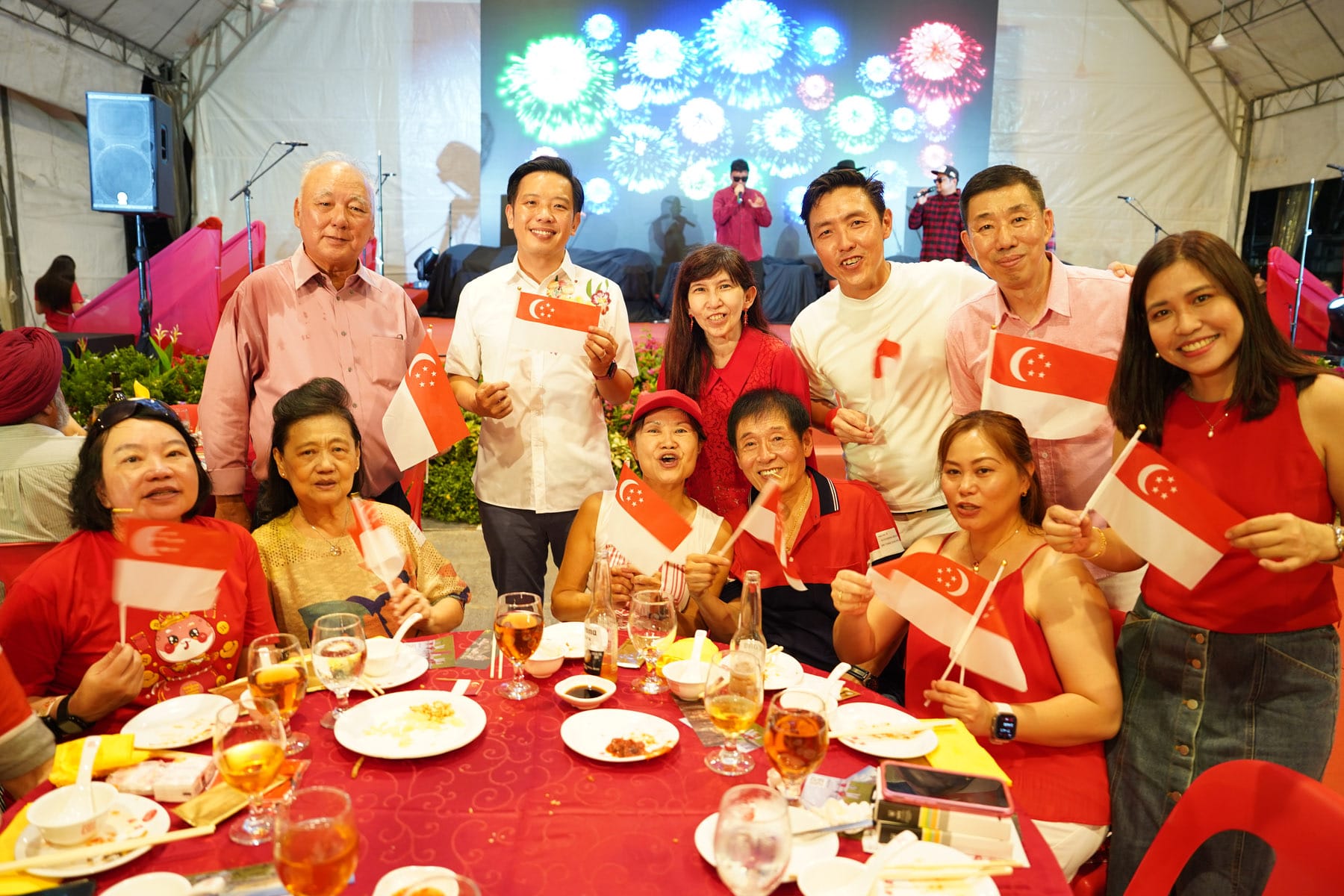 Group of people smiling and holding Singapore flags at a festive dinner event with colorful fireworks display on screen behind them.