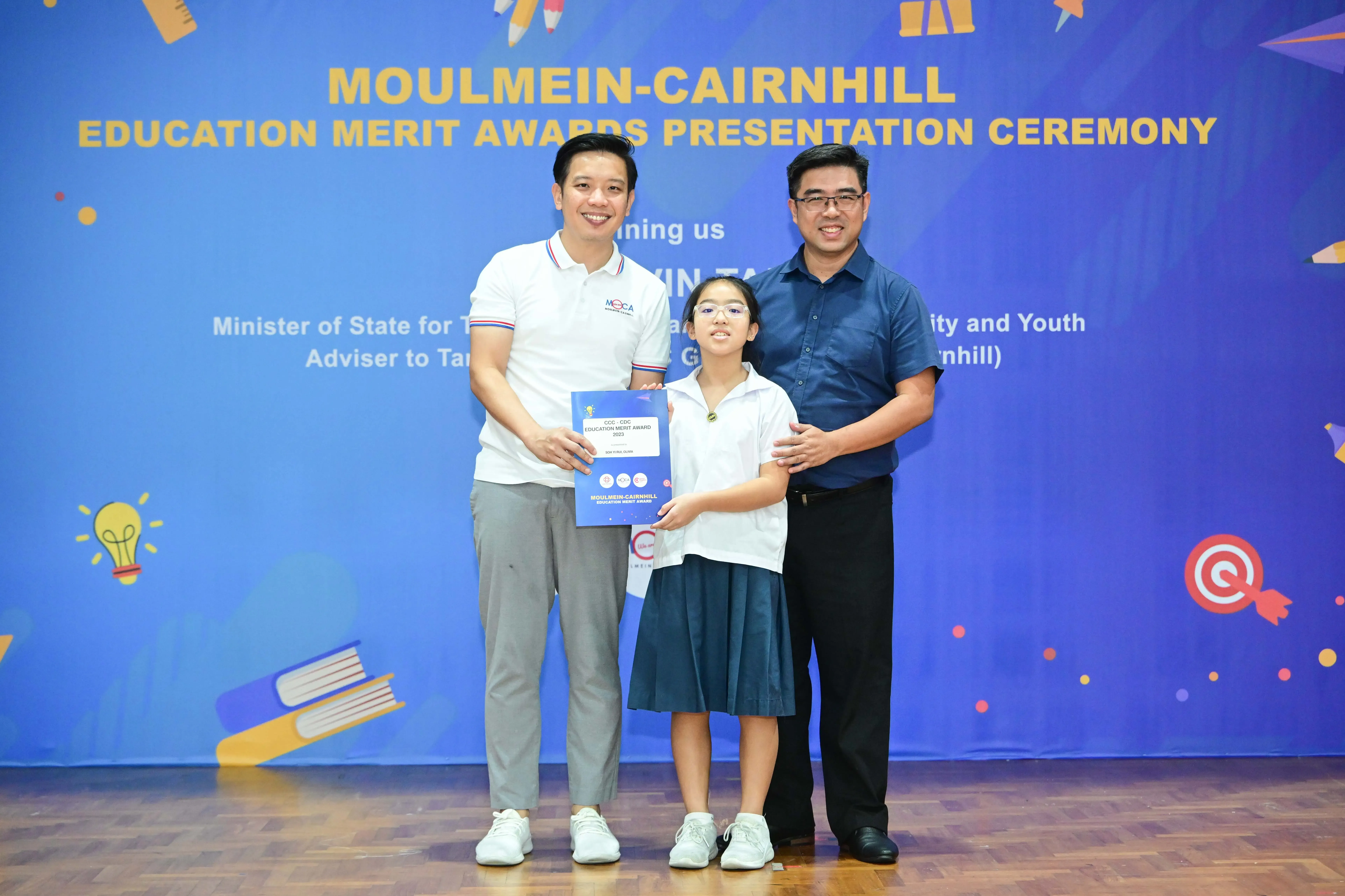 Two men and a young girl in school uniform hold a certificate at the Moulmein-Cairnhill Education Merit Awards ceremony.