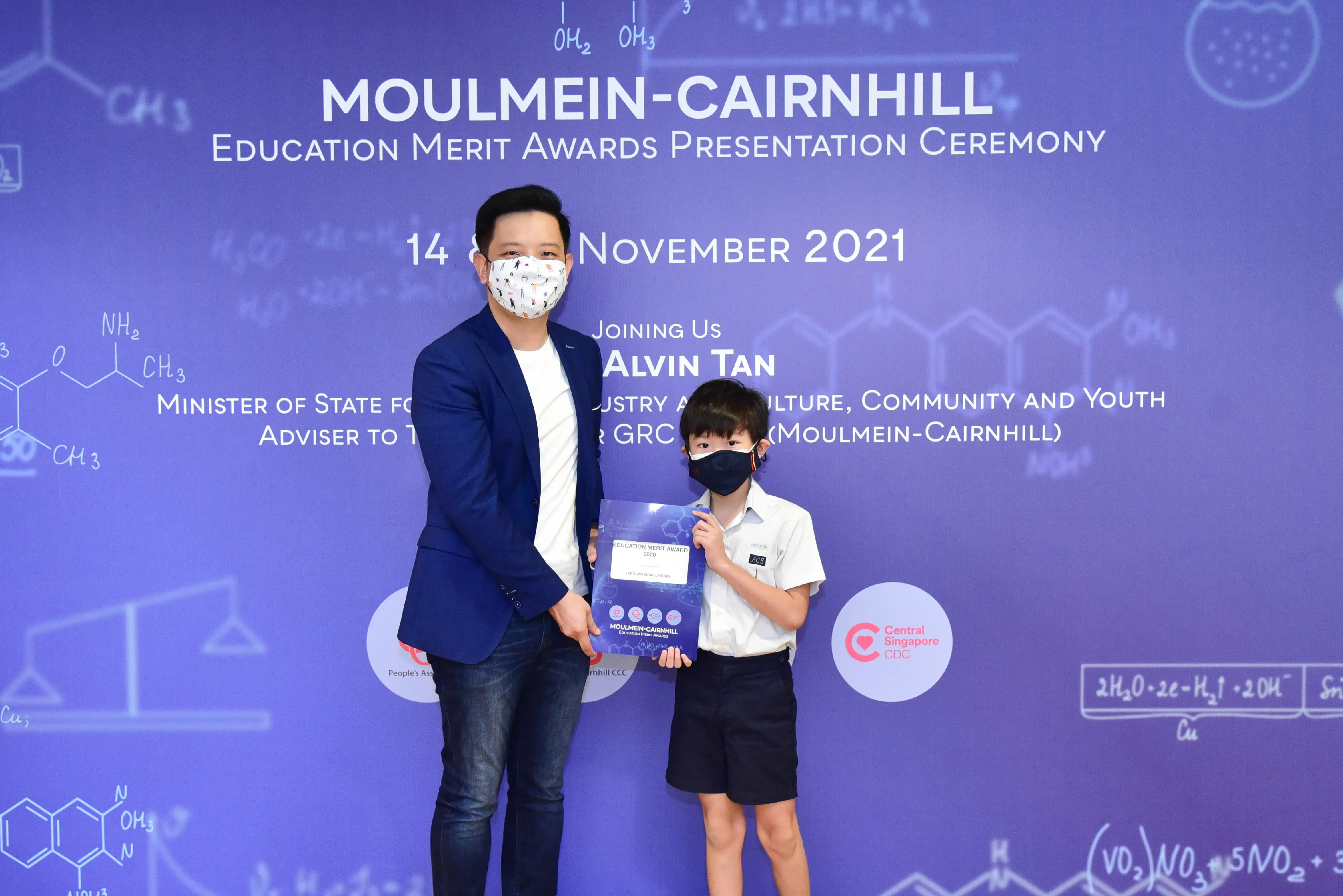 An adult and a young boy in masks hold an award certificate at the Moulmein-Cairnhill Education Merit Awards ceremony.