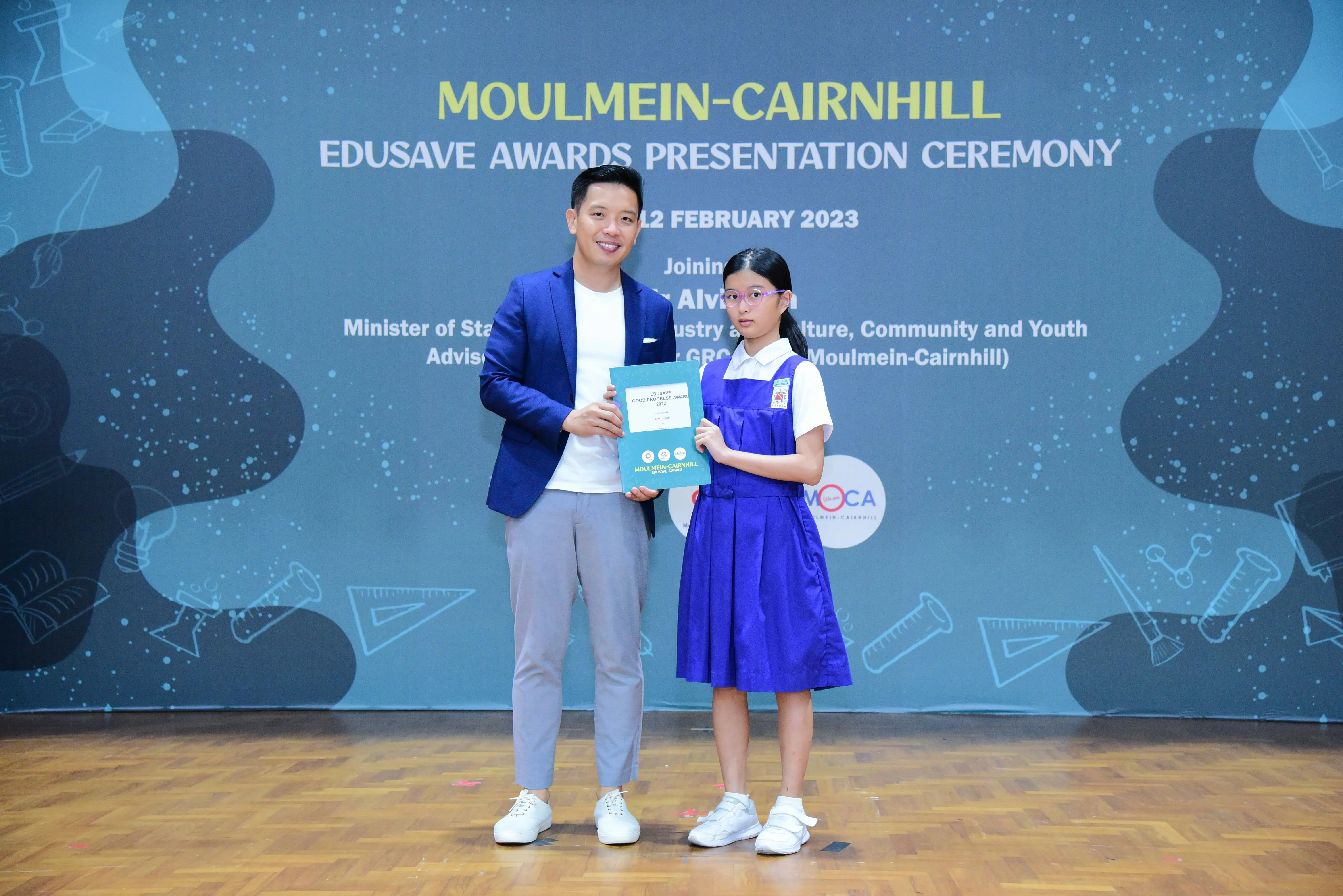 A man in a blue blazer and a young girl in a blue school uniform hold a certificate at the Moulmein-Cairnhill Edusave Awards 2023.