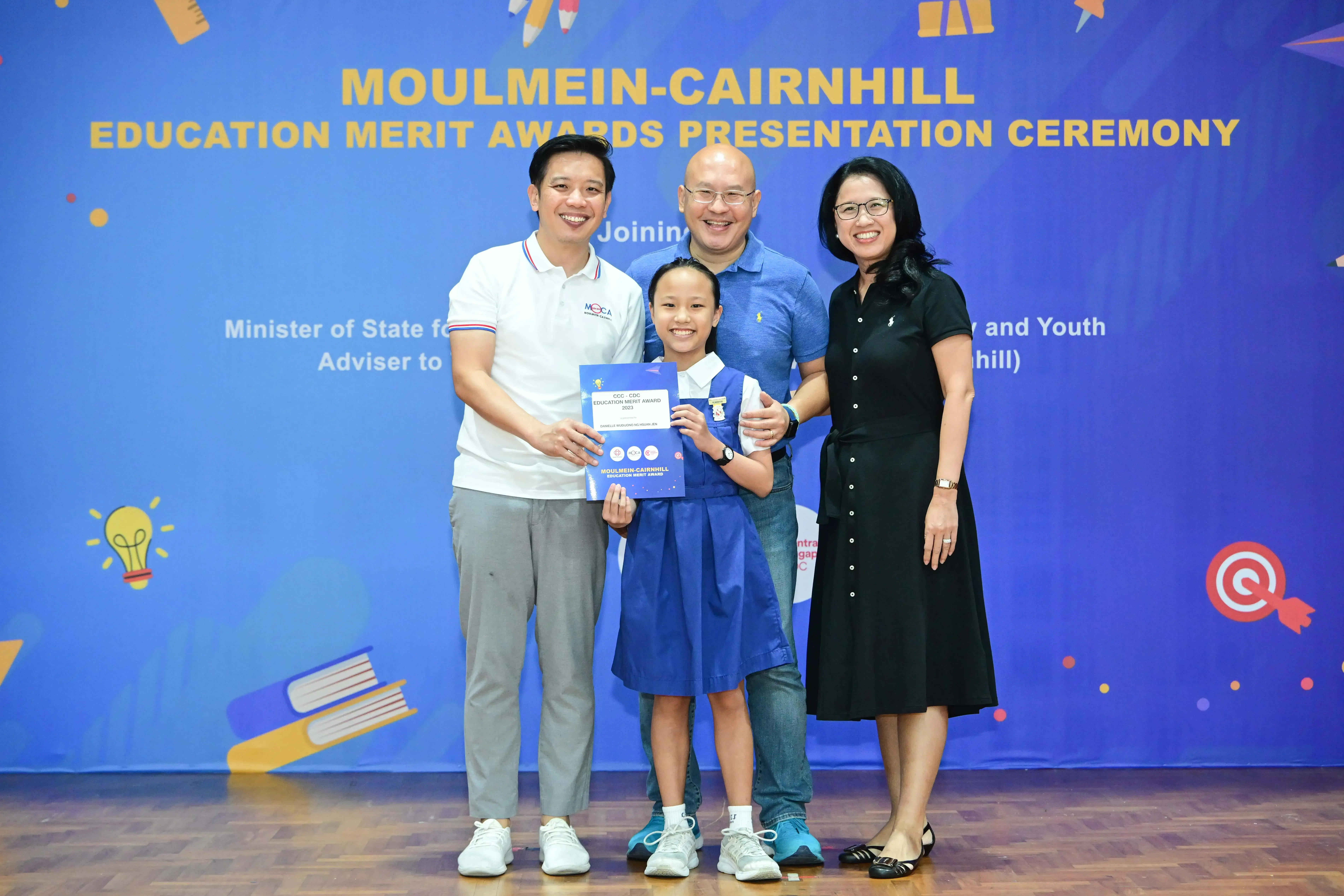 A young girl in a blue uniform holds an award certificate alongside three smiling adults at the Moulmein-Cairnhill Education Merit Awards ceremony.