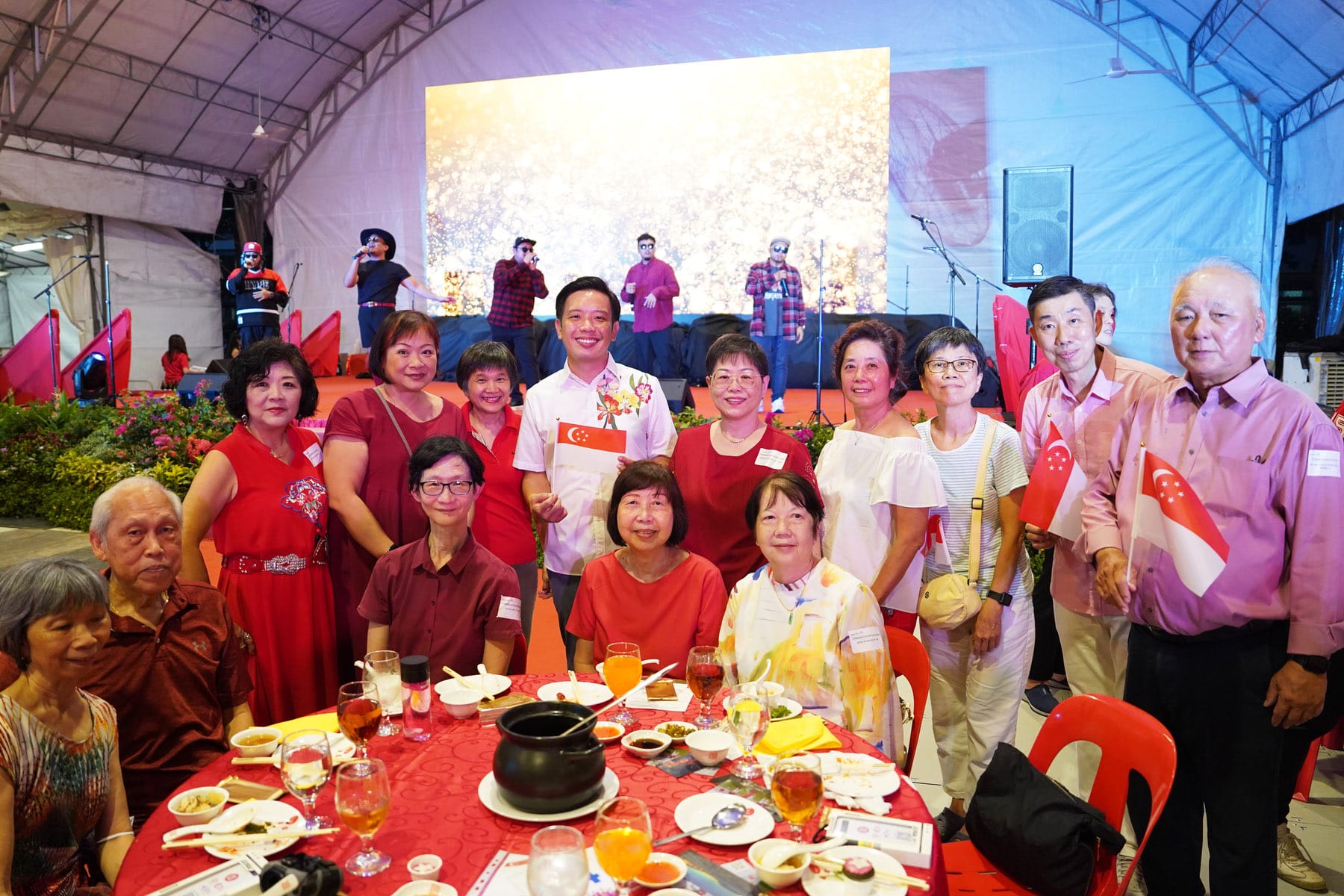 A group of people in red attire pose at a dinner table, holding Singapore flags, with performers on a stage behind them.