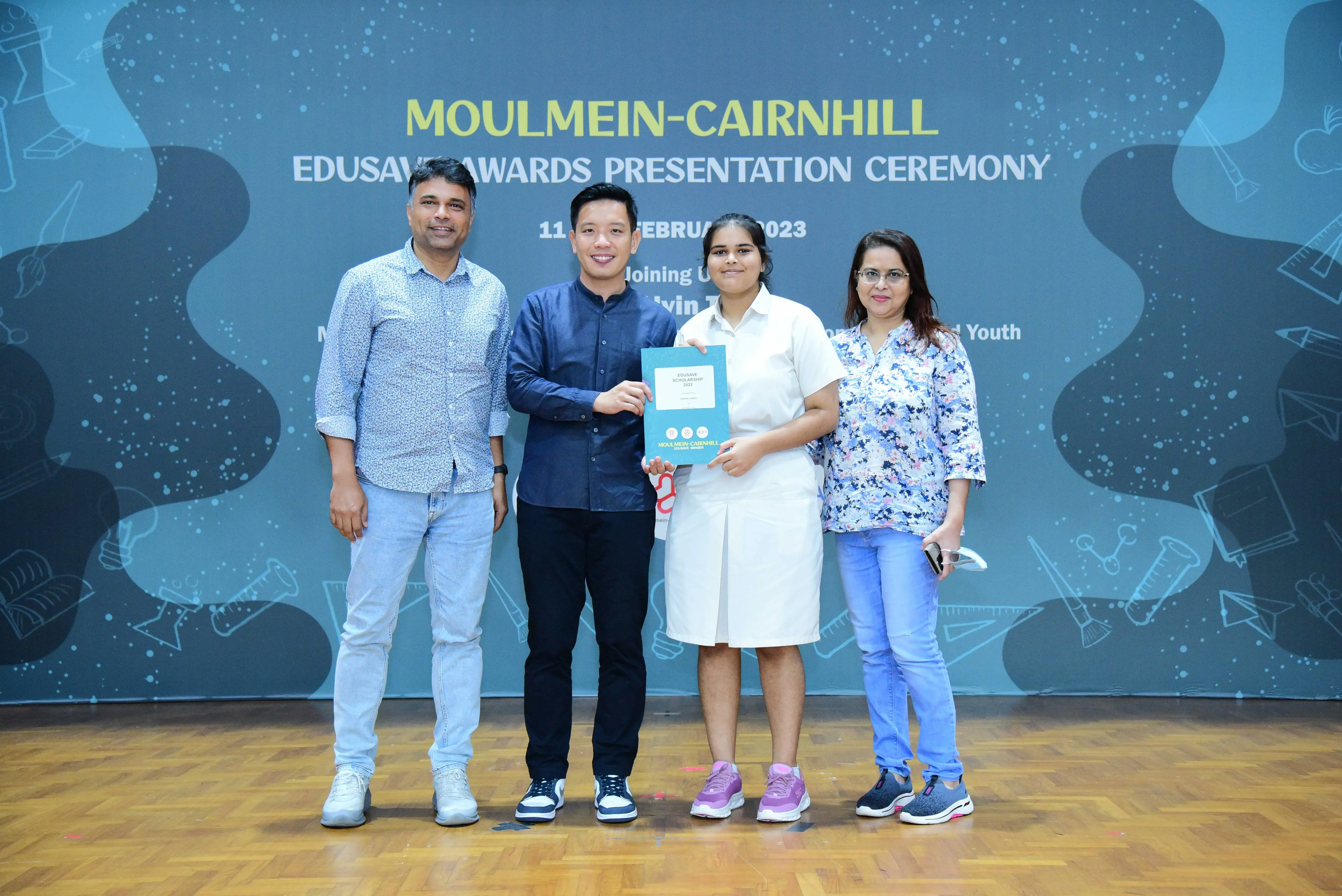 Four people pose on a stage at the Moulmein-Cairnhill Edusave Awards Presentation Ceremony 2023, holding a certificate.