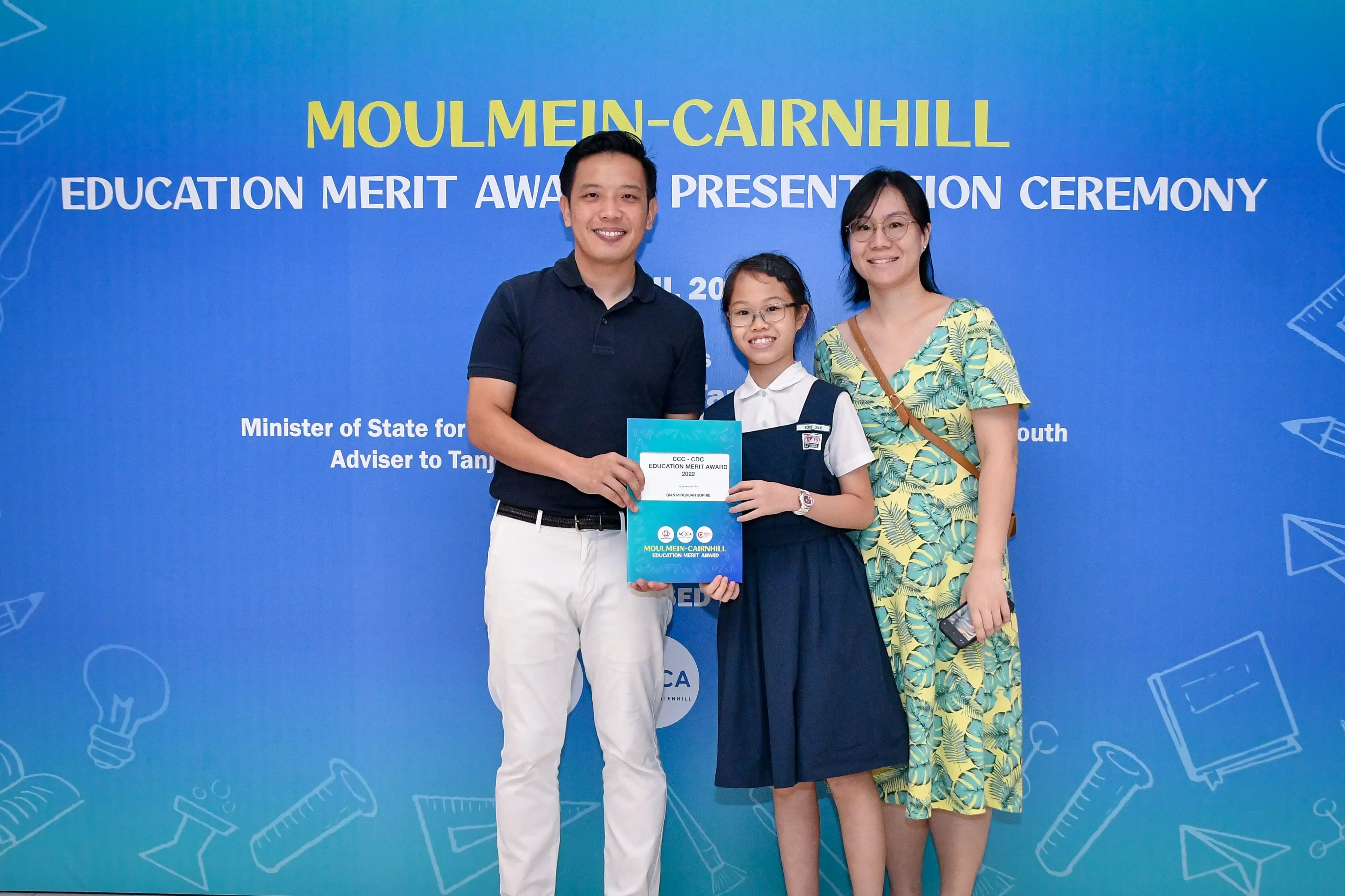 A girl holding an award certificate stands between two adults at the Moulmein-Cairnhill Education Merit Award Presentation Ceremony.