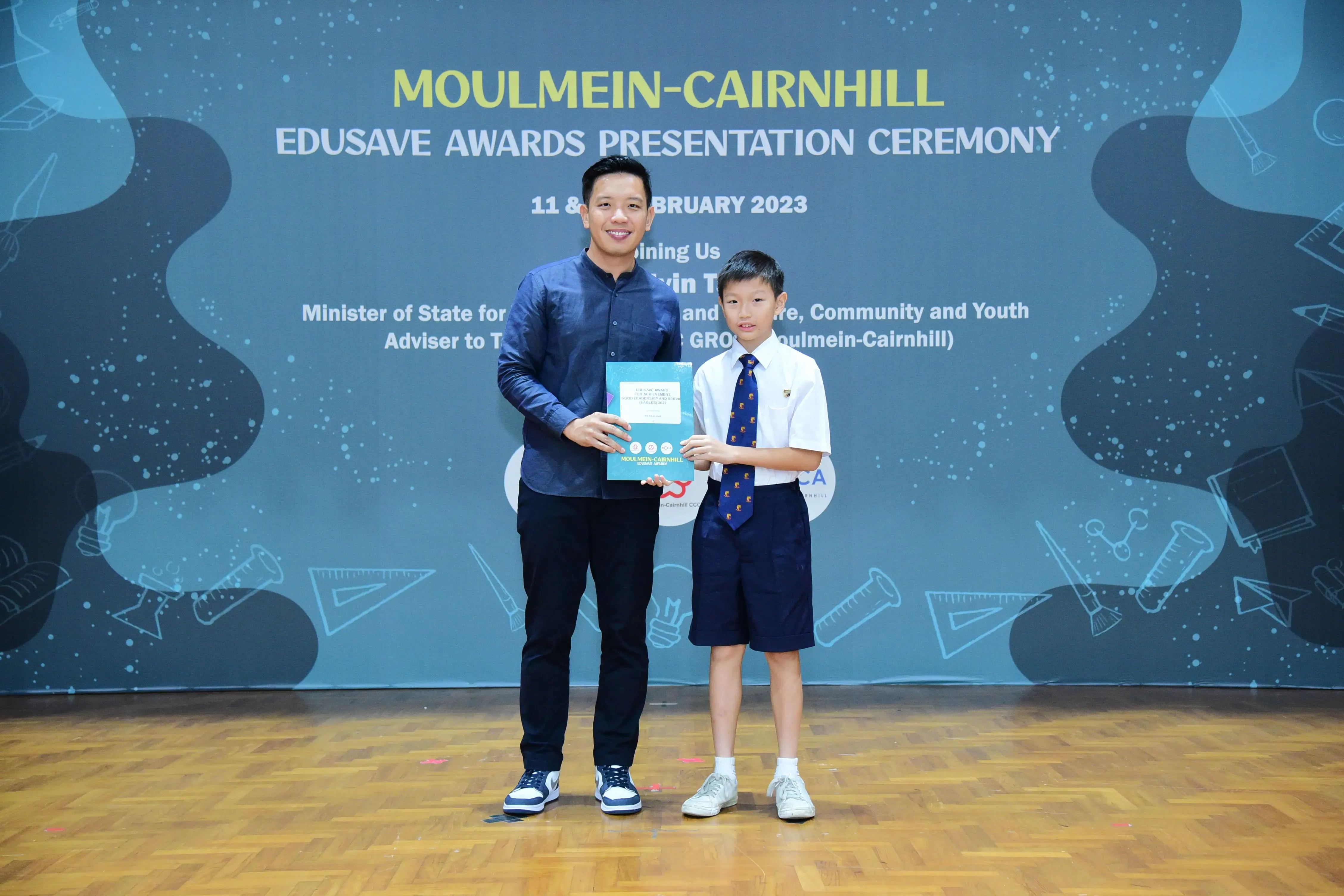 An adult and a young student in uniform hold a certificate at the Moulmein-Cairnhill Edusave Awards Ceremony 2023.