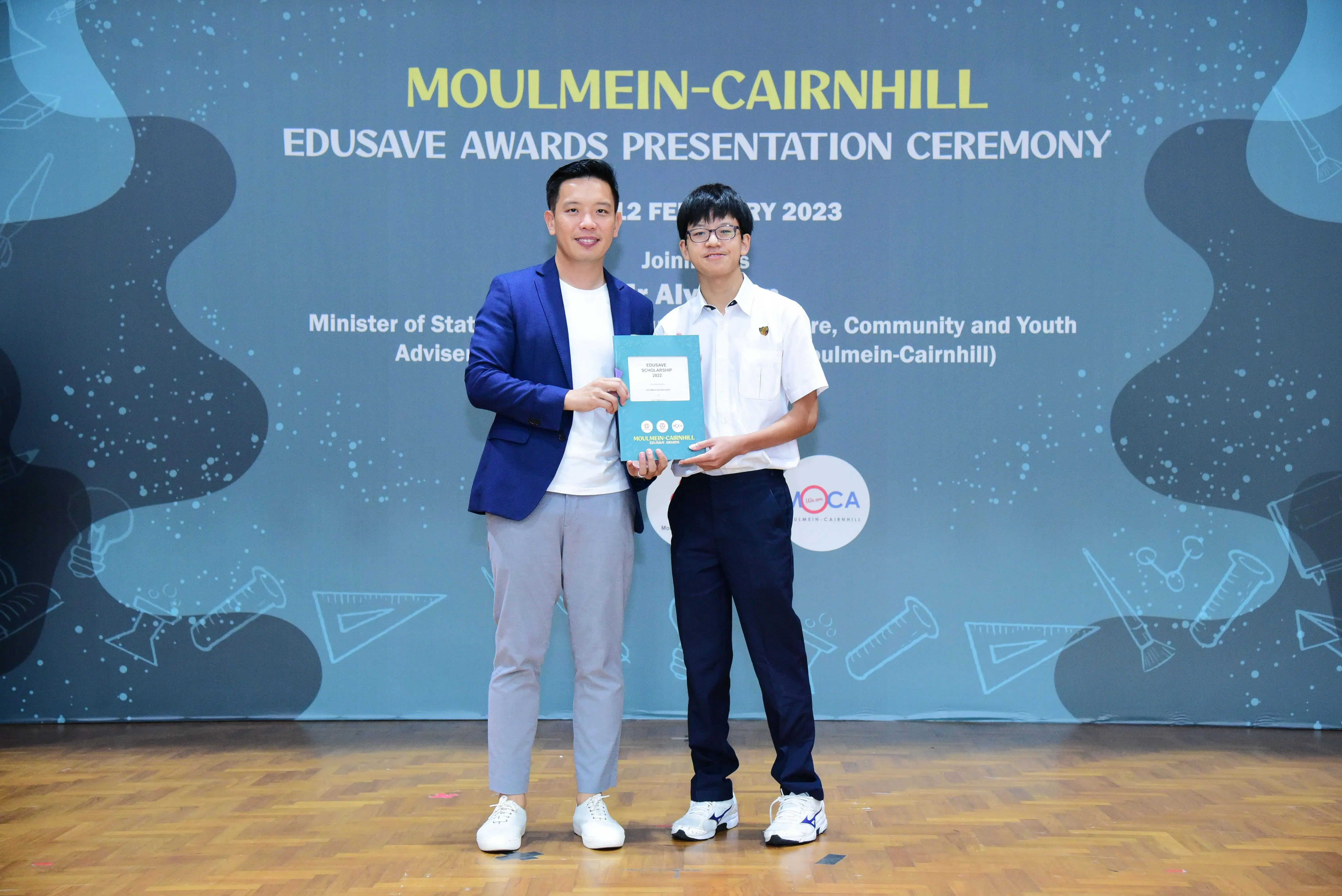 Two men hold a certificate together at the Moulmein-Cairnhill Edusave Awards Presentation Ceremony, February 2023.