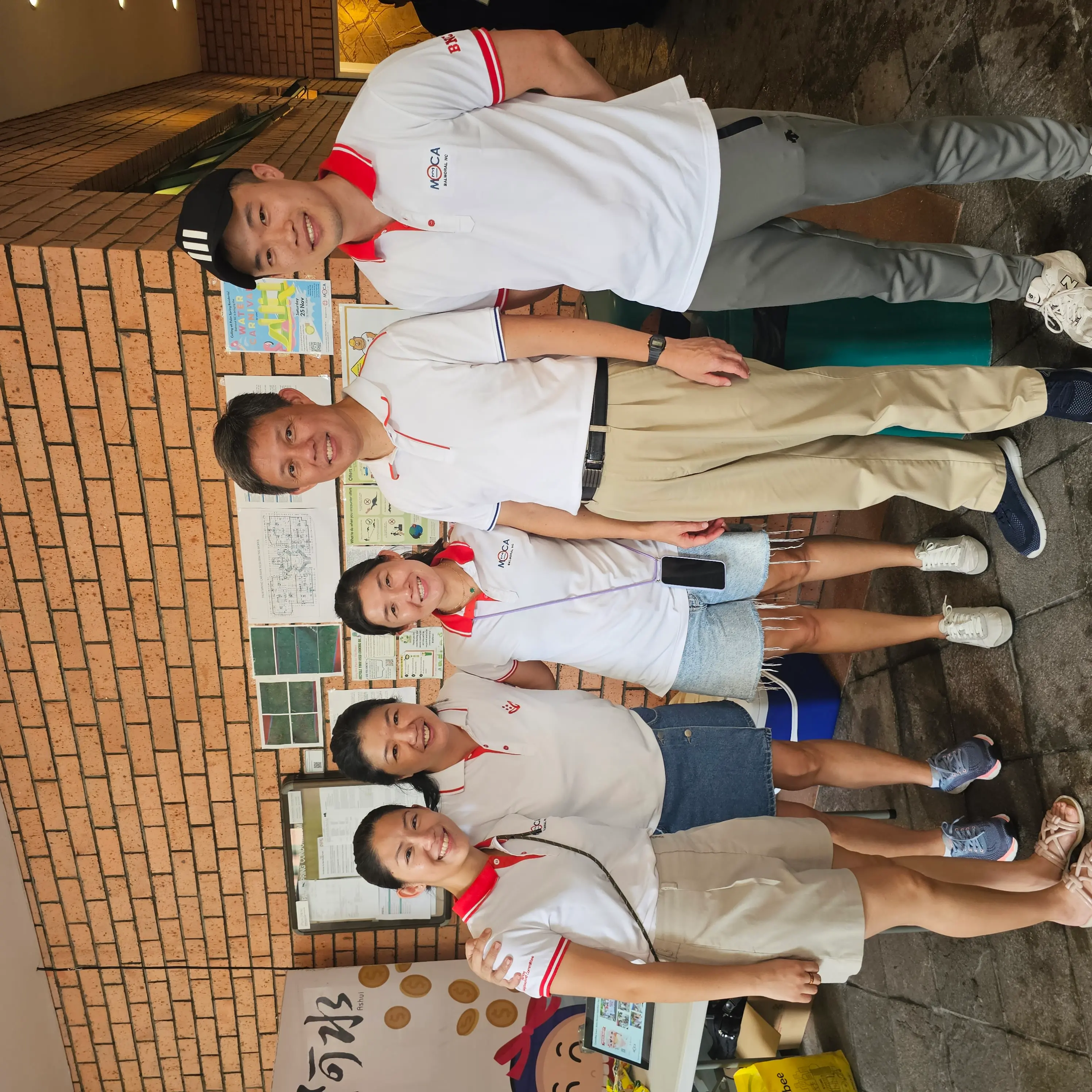 Five people in white polo shirts smile together outdoors near a brick wall with informational posters displayed.