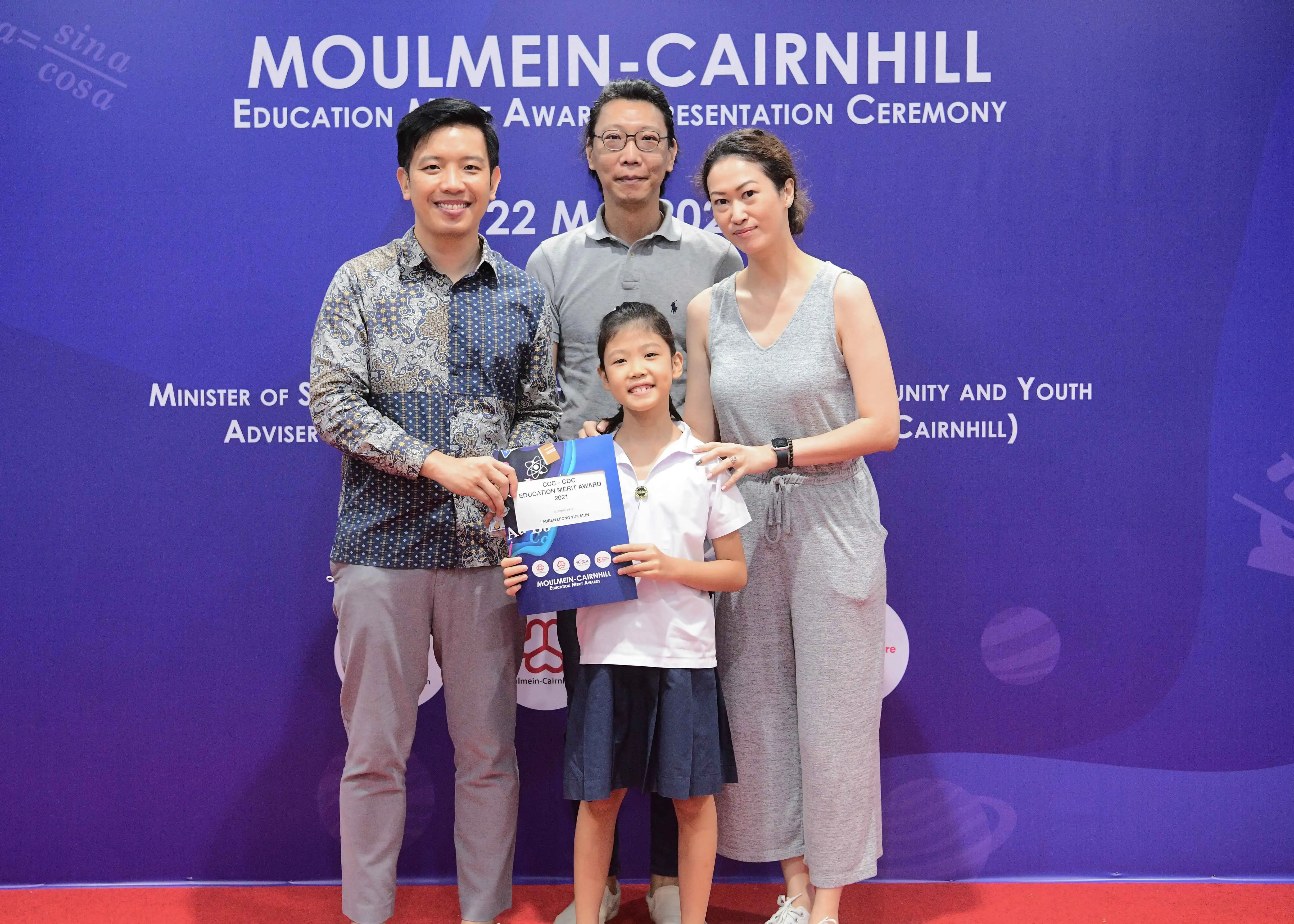 Four people pose at the Moulmein-Cairnhill Education Merit Award ceremony, with a young girl holding an award folder.