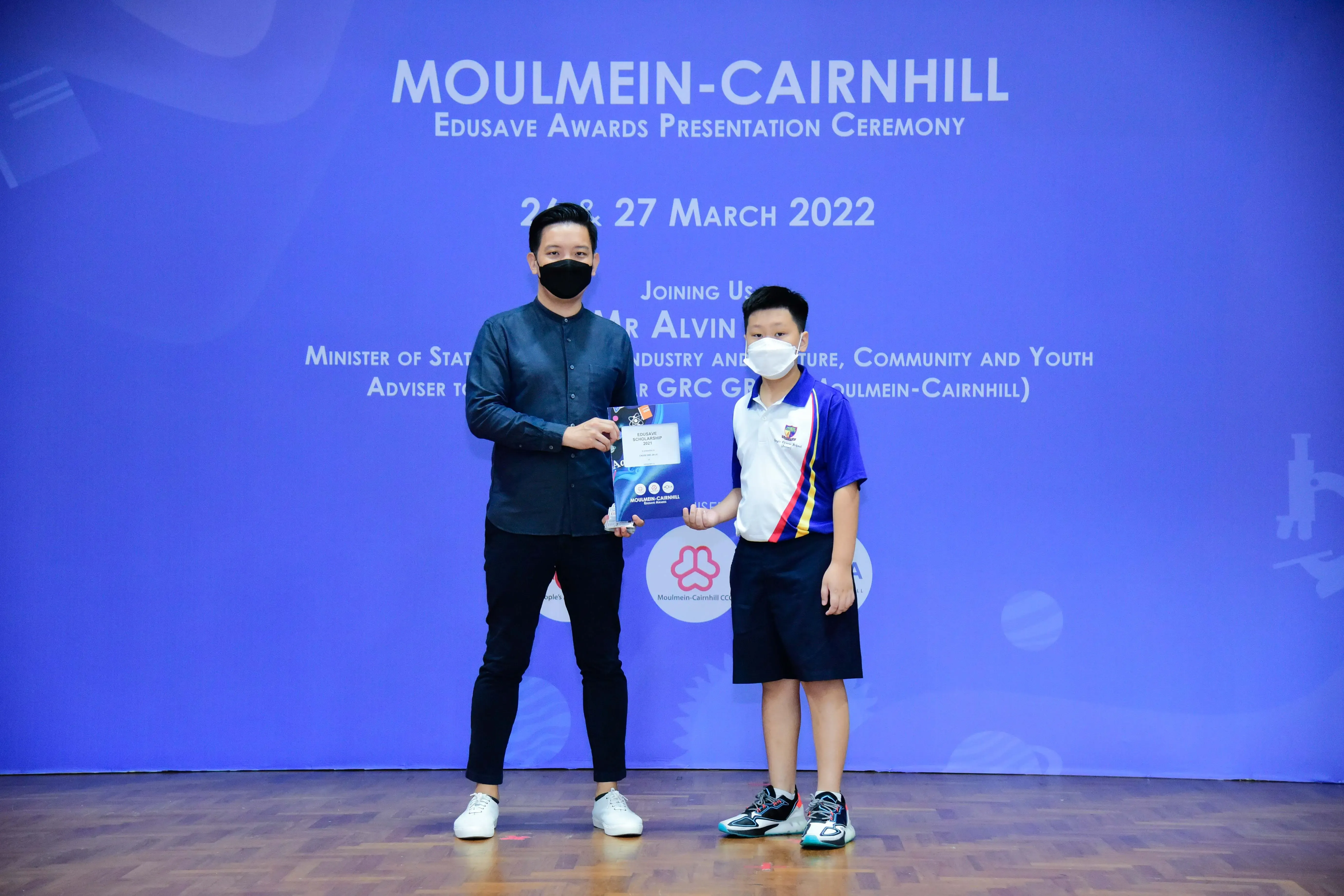 An adult and a young student in school uniform hold an award together at the Moulmein-Cairnhill Edusave ceremony, March 2022.