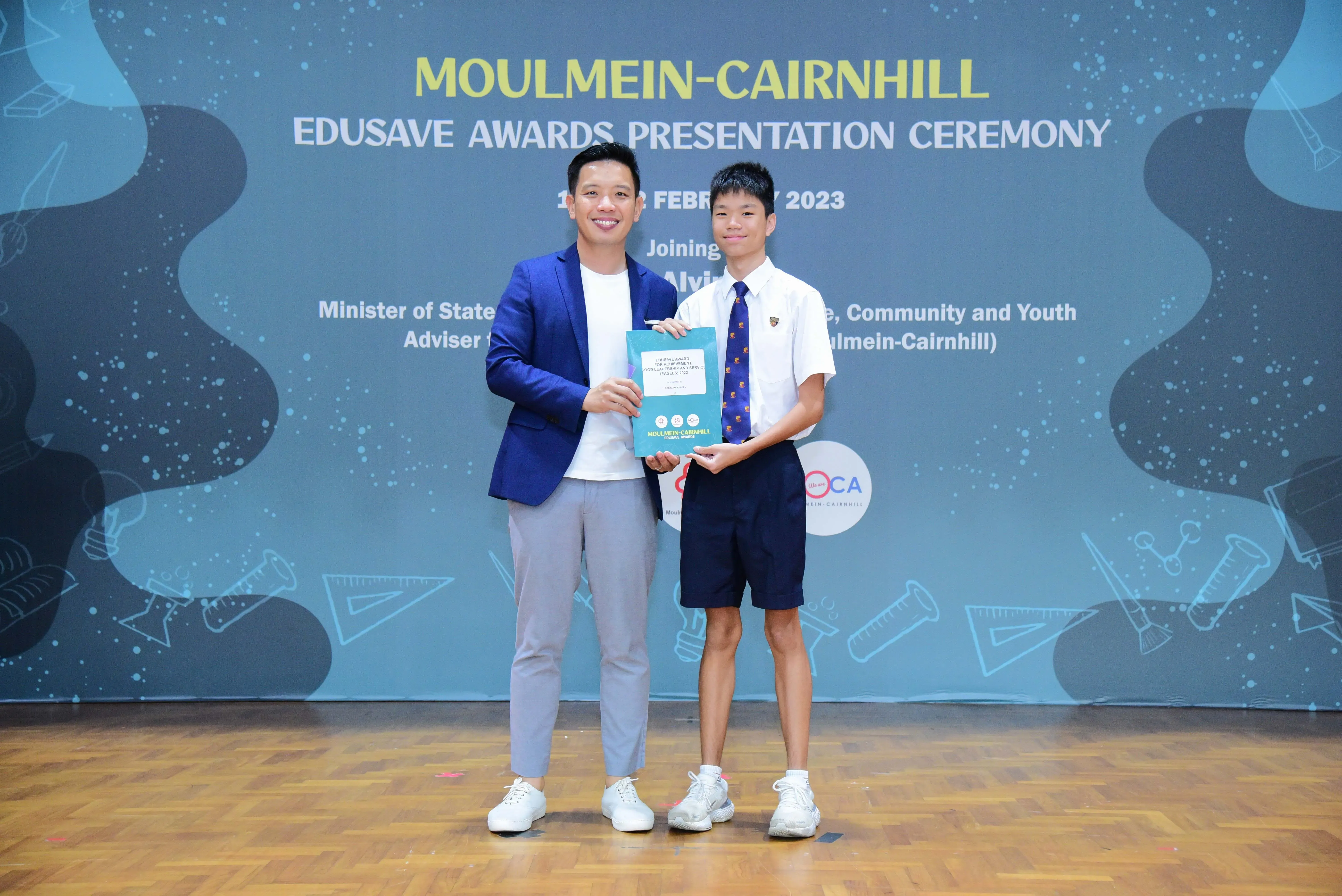An adult in a blue blazer and a uniformed student hold a certificate at the Moulmein-Cairnhill Edusave Awards 2023.