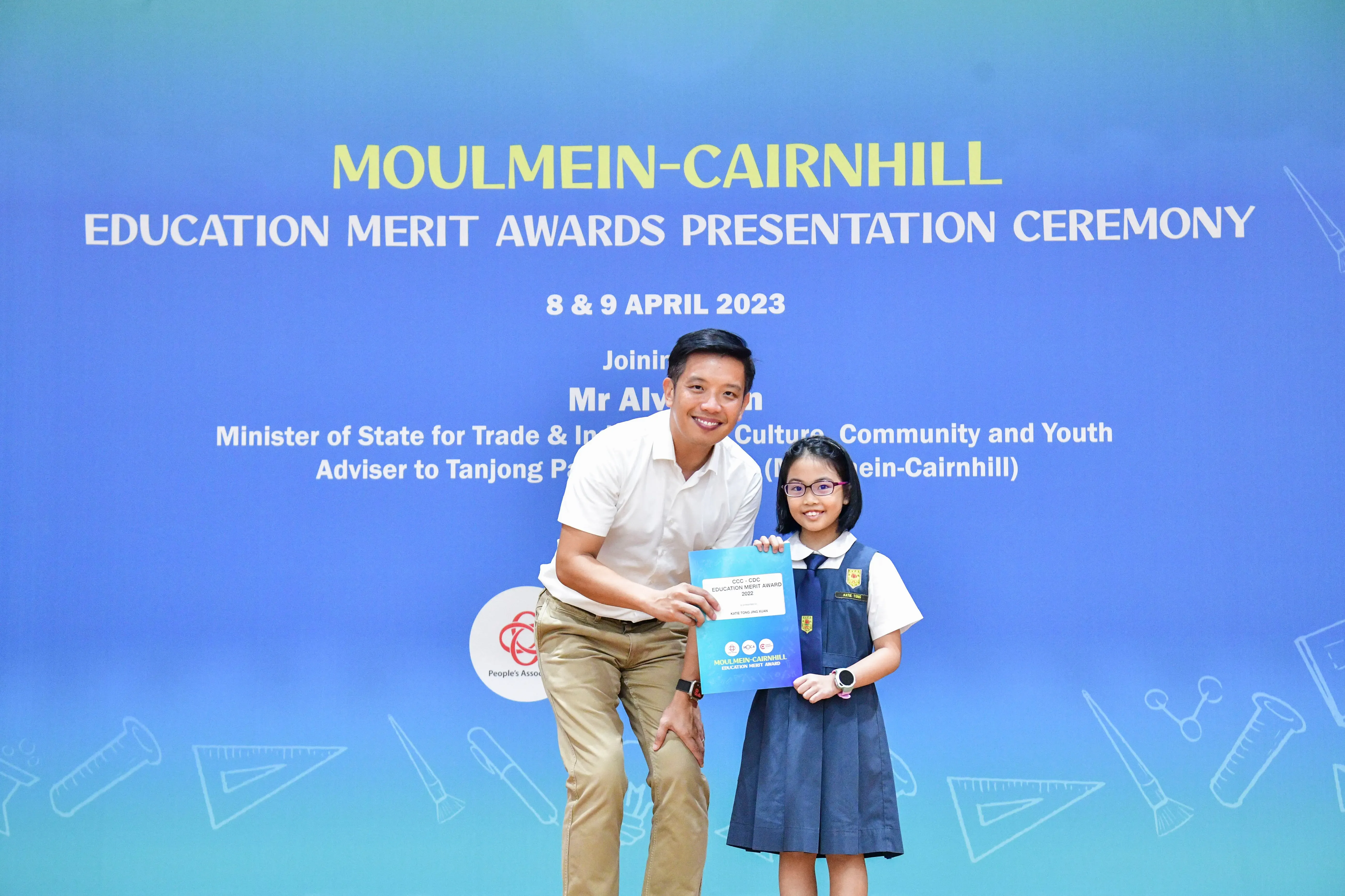 A man and a young girl in school uniform hold an award certificate in front of a Moulmein-Cairnhill Education Merit Awards banner.