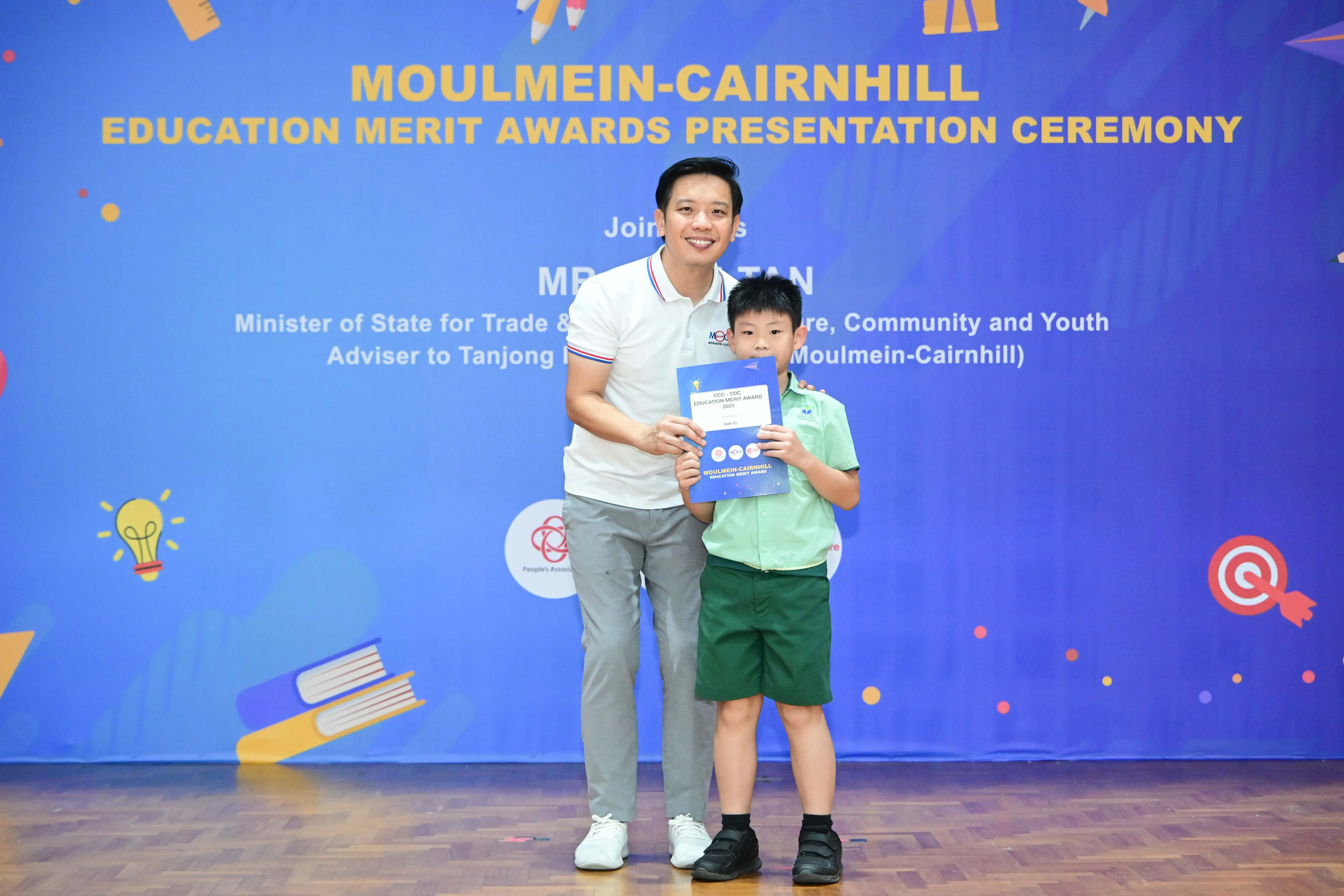 A man and a young boy hold a certificate together in front of a Moulmein-Cairnhill Education Merit Awards banner.