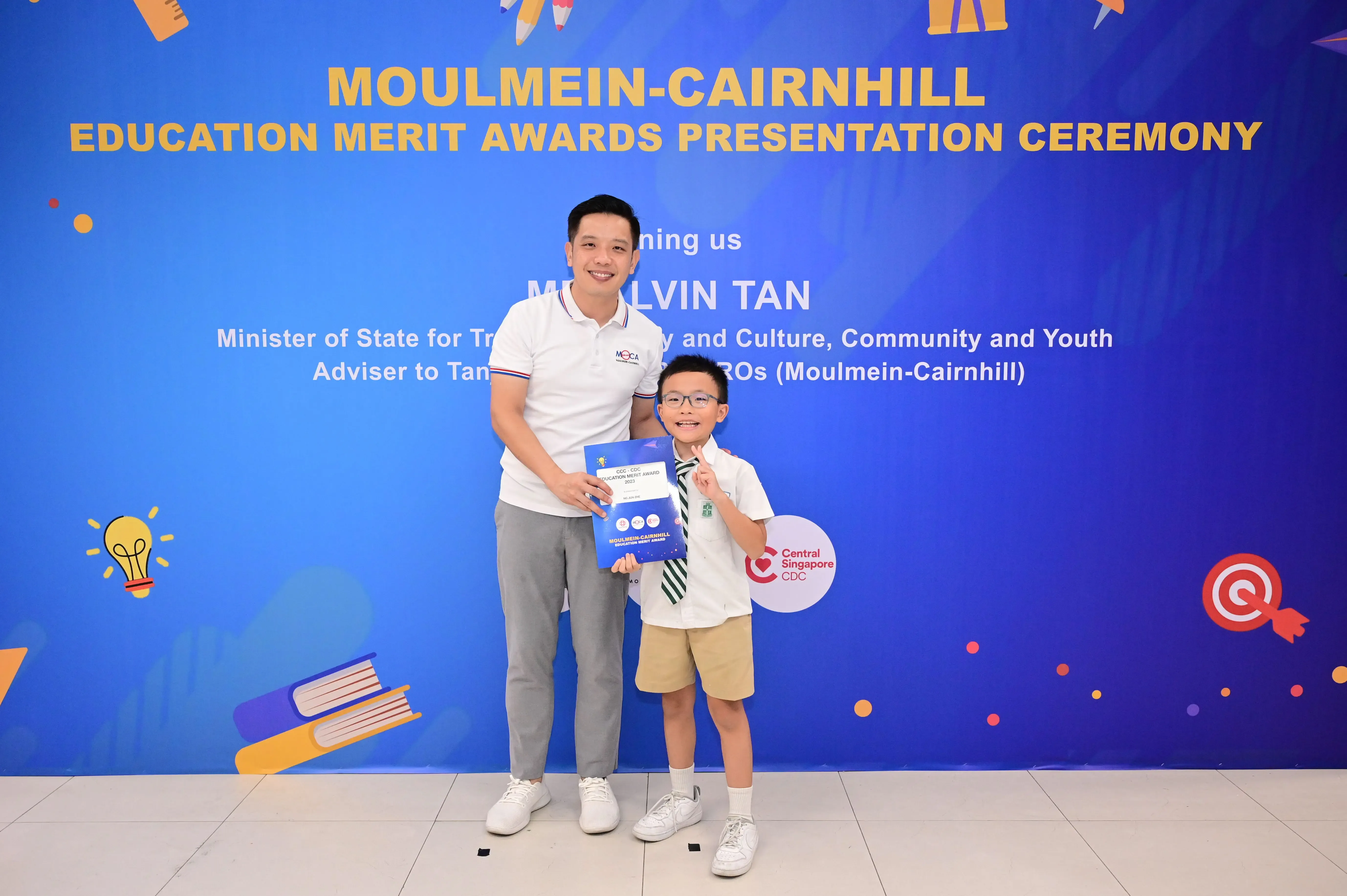 A man and a young boy holding an award certificate stand before a Moulmein-Cairnhill Education Merit Awards banner.