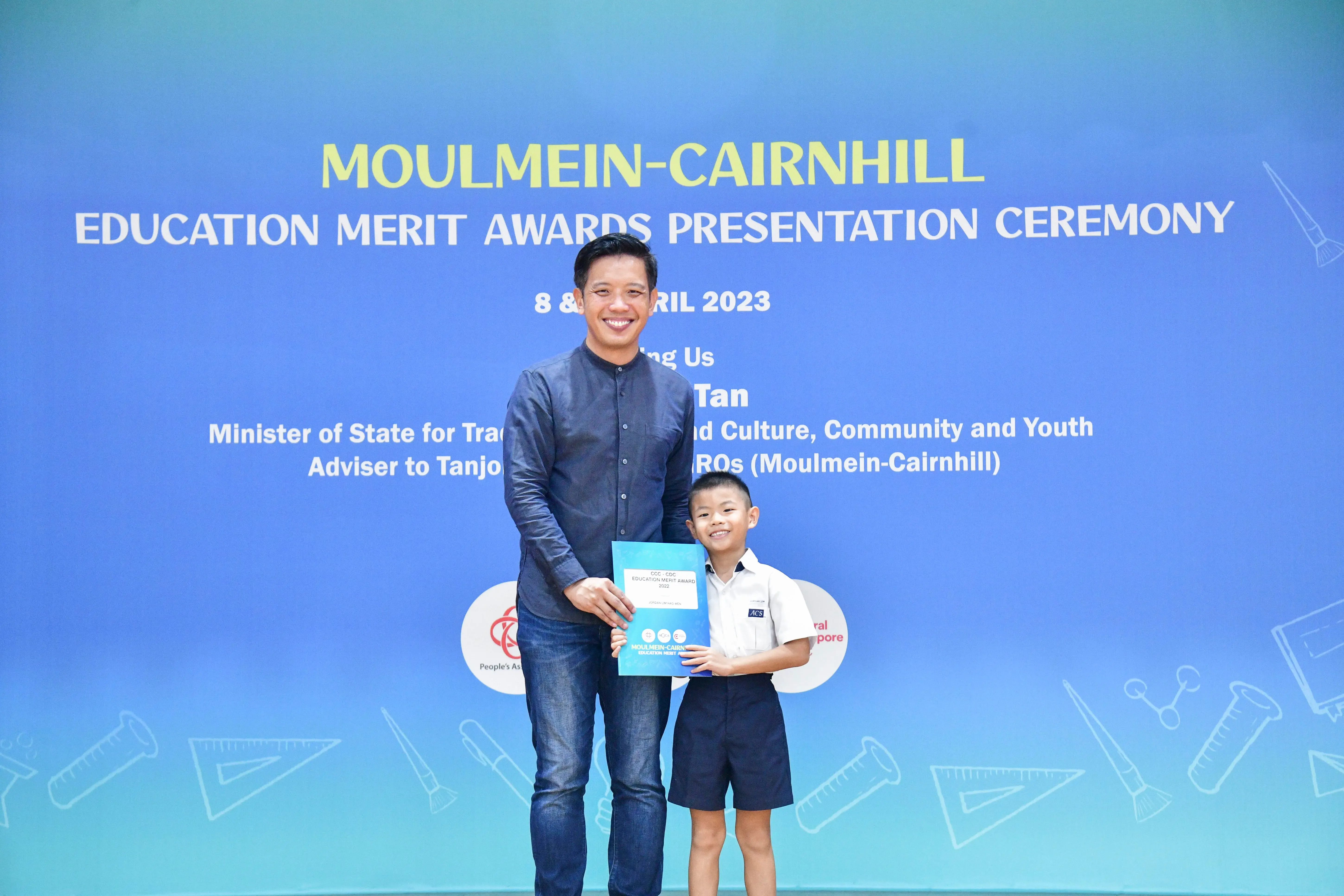 A man and a young boy holding a certificate stand before a blue Moulmein-Cairnhill Education Merit Awards banner.