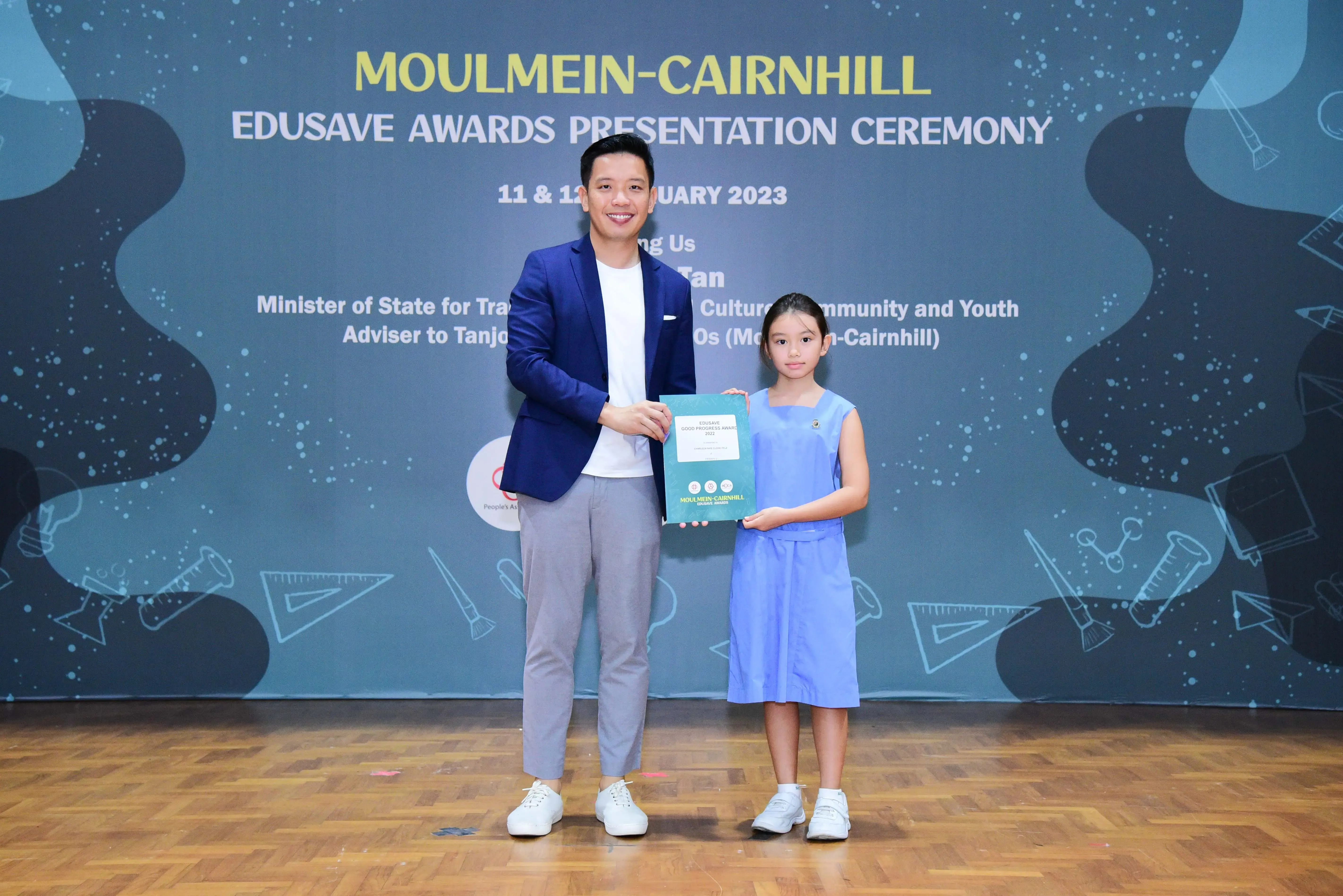 A man in a blue blazer and a young girl in a blue dress hold an award at the Moulmein-Cairnhill Edusave ceremony 2023.