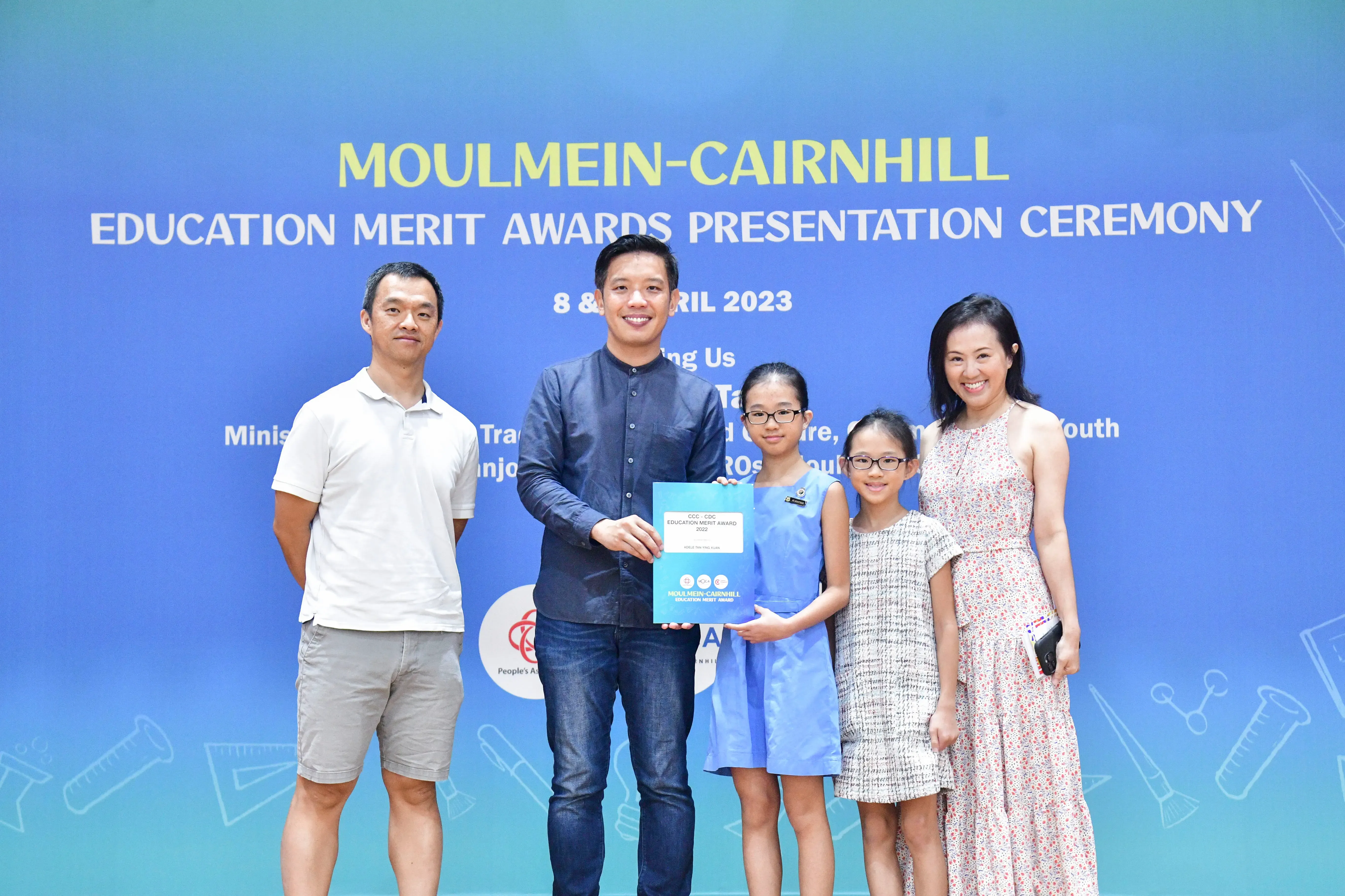 Five people pose before a Moulmein-Cairnhill Education Merit Awards Presentation Ceremony banner, holding a blue award certificate.