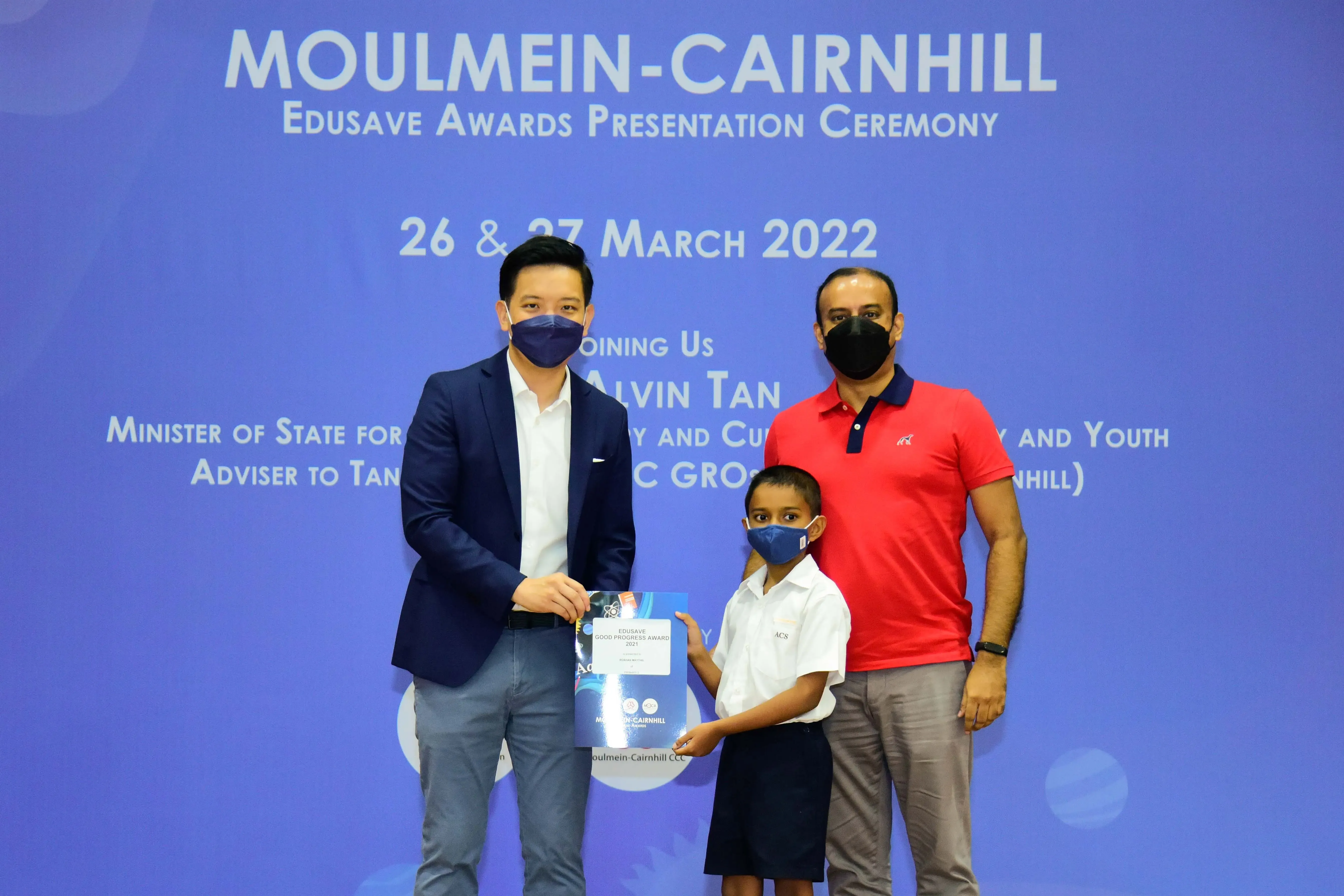 Two masked adults and a masked child hold a certificate at the Moulmein-Cairnhill Edusave Awards Ceremony, March 2022.
