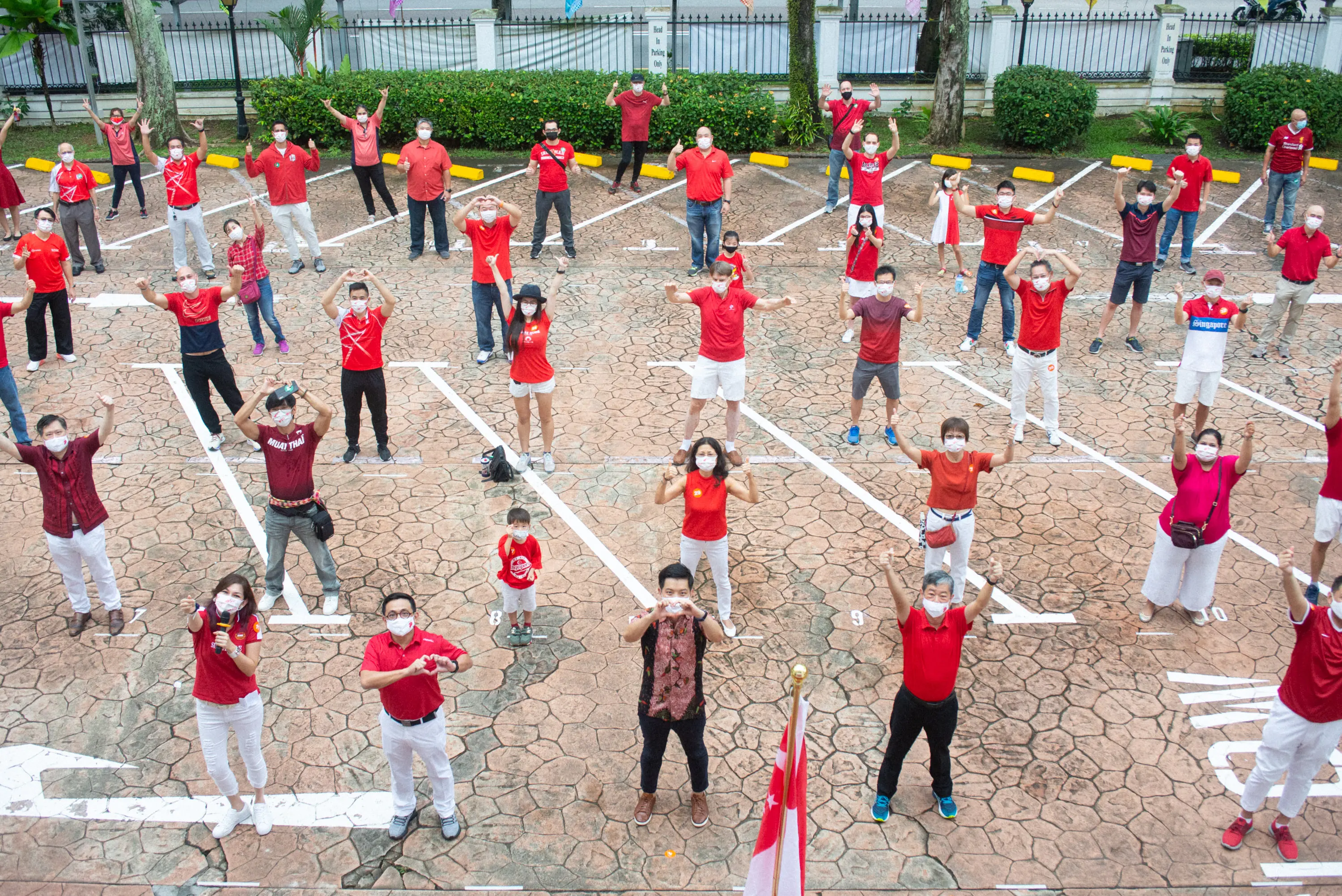 Large group of masked people in red clothing standing spaced apart in a parking lot, forming heart shapes with their hands.