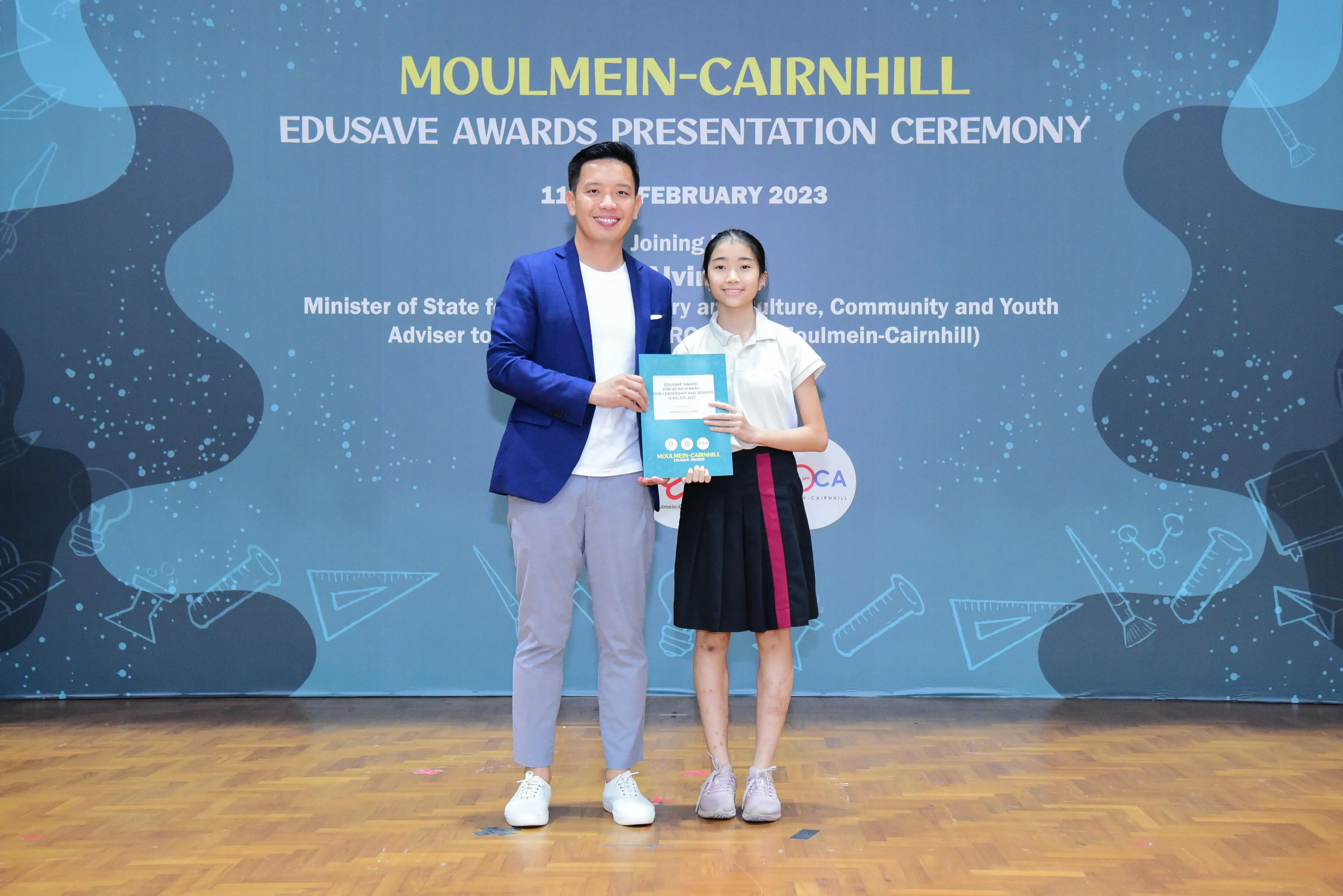 A man in a blue blazer and a young girl in school uniform hold an award at the Moulmein-Cairnhill Edusave Awards 2023.