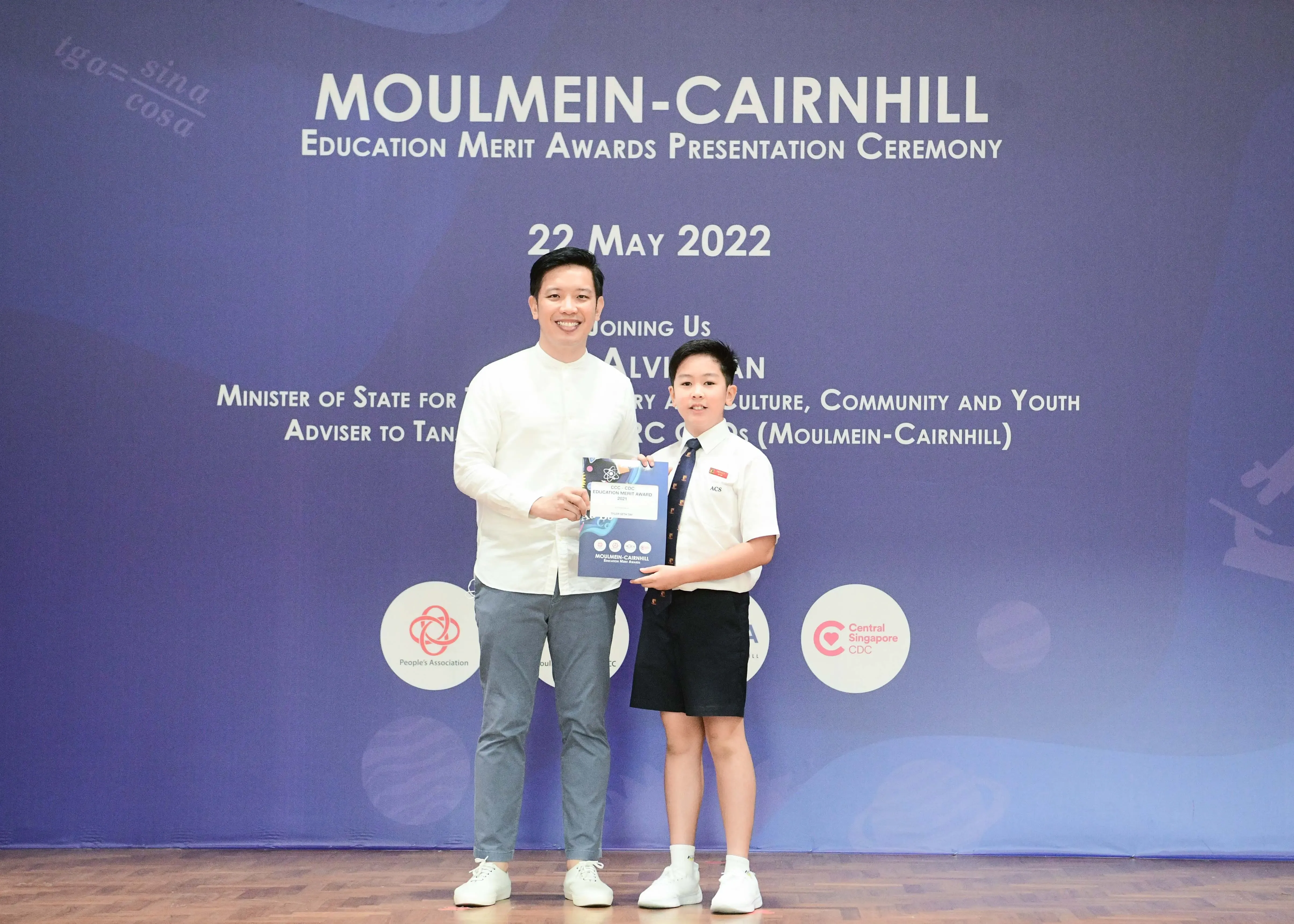 A man and a young boy in school uniform hold an award certificate at the Moulmein-Cairnhill Education Merit Awards ceremony.