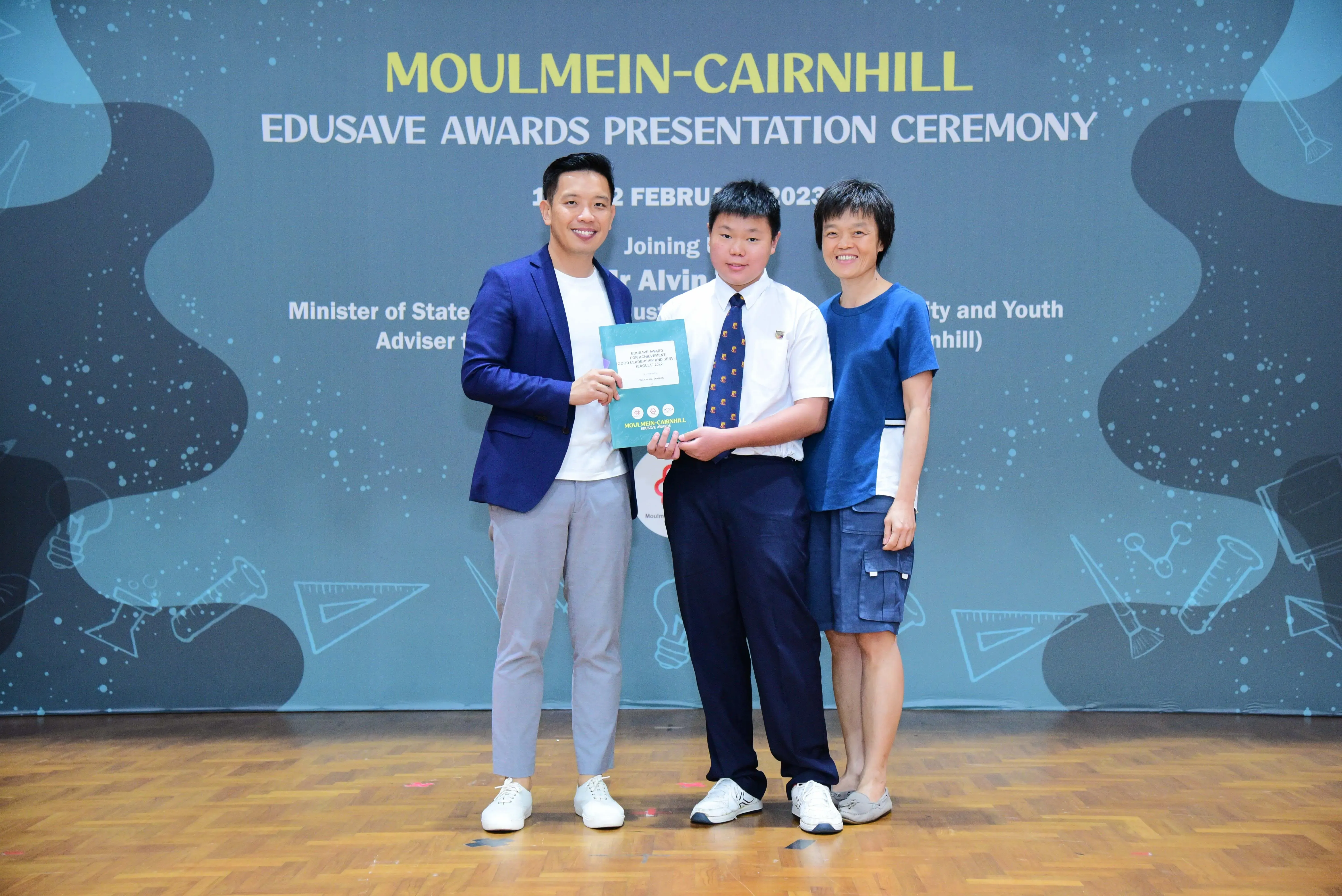 Three people pose on a stage holding an award booklet at the Moulmein-Cairnhill Edusave Awards Presentation Ceremony.