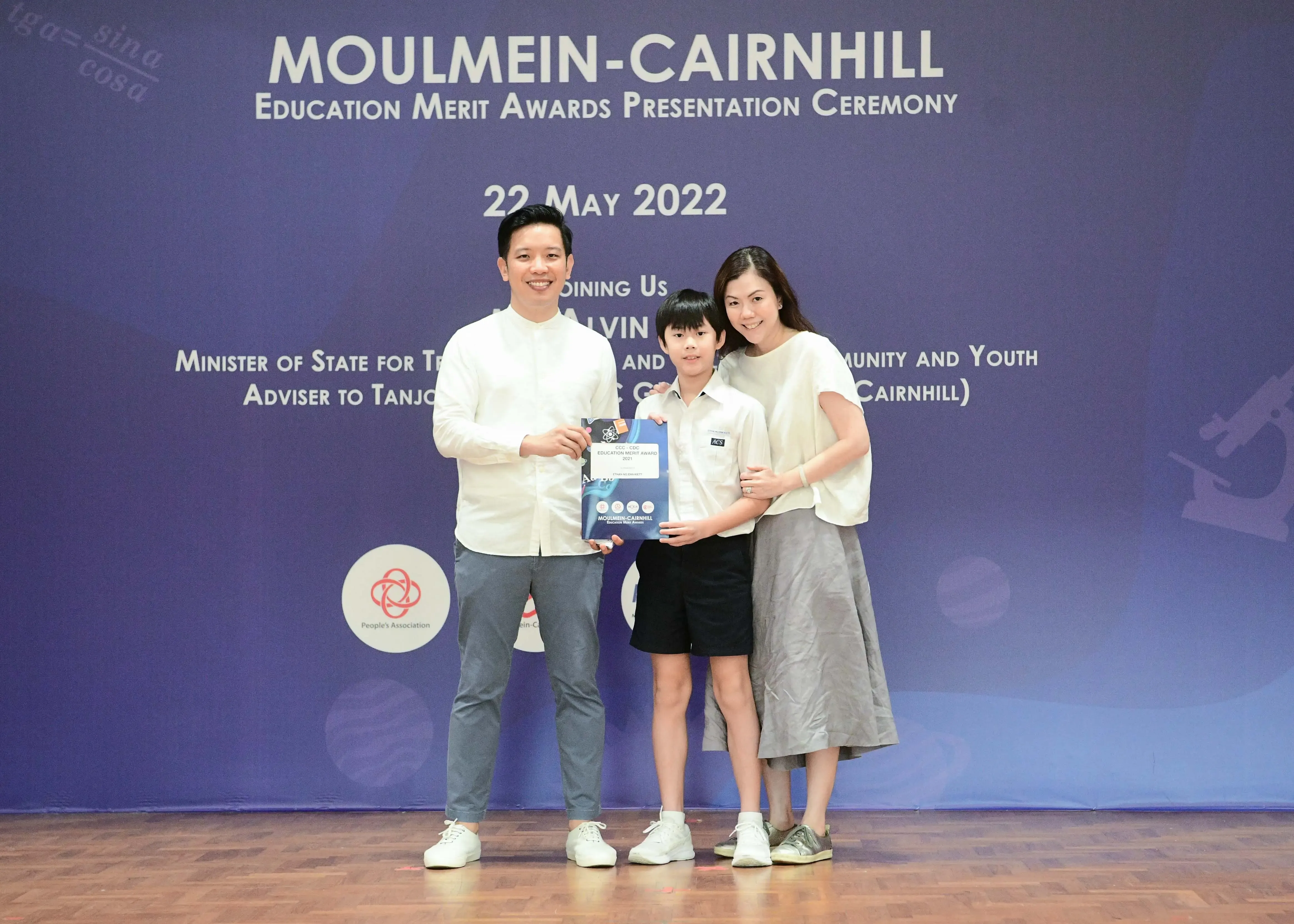 A boy holding an award certificate stands between two smiling adults at the Moulmein-Cairnhill Education Merit Awards ceremony.