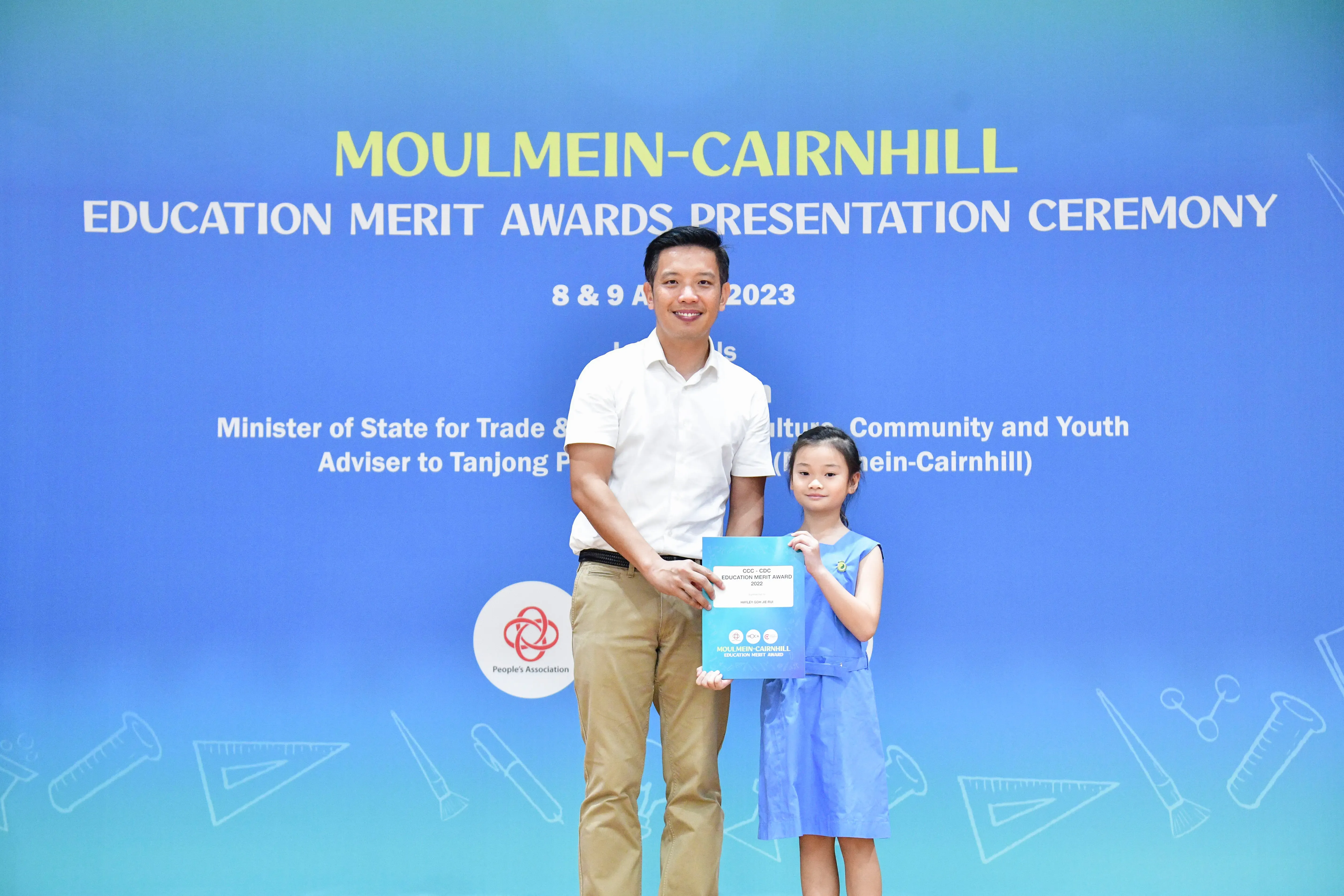 Man and young girl hold an award certificate in front of a Moulmein-Cairnhill Education Merit Awards 2023 banner.