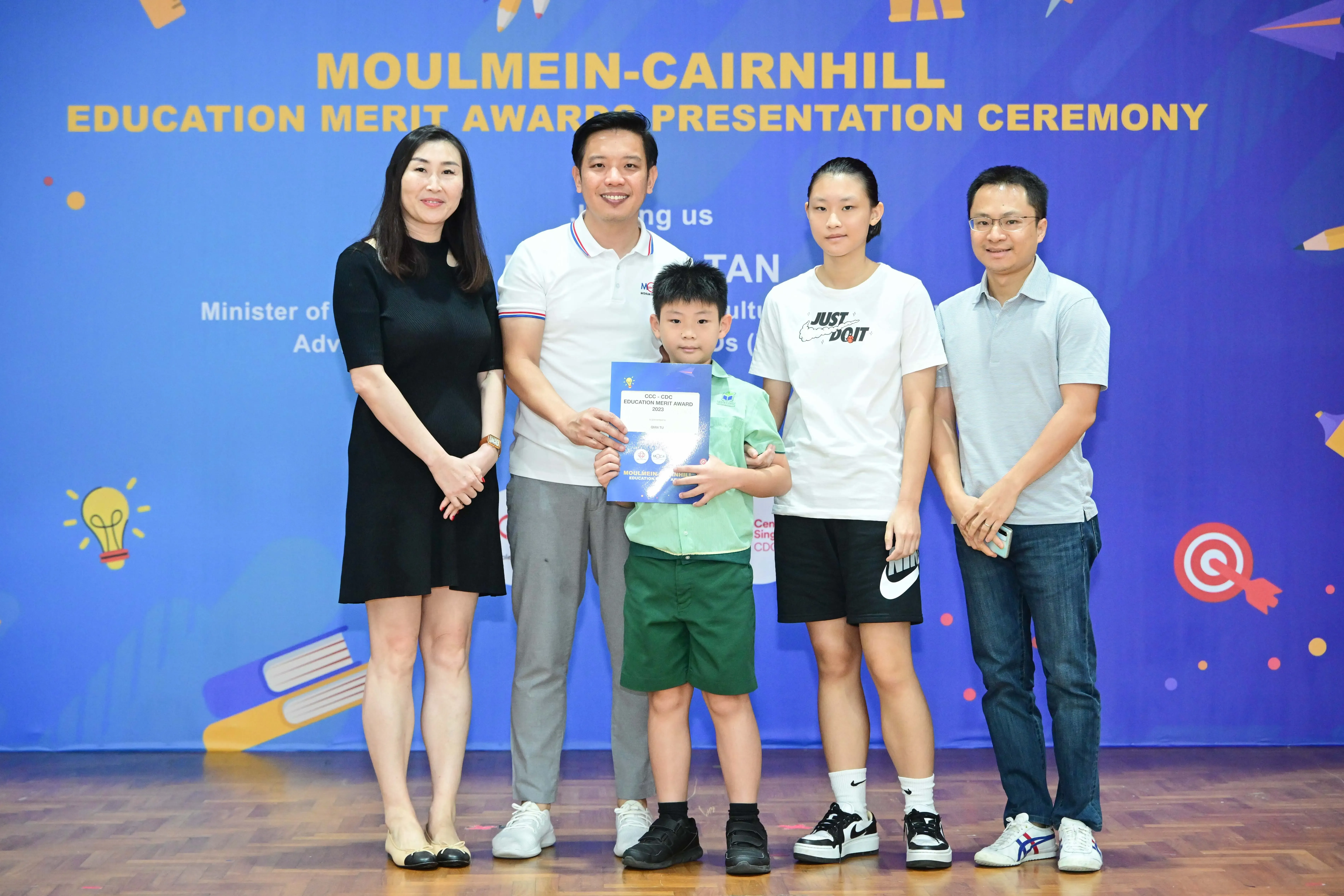 Five people pose at the Moulmein-Cairnhill Education Merit Awards Ceremony, with a young boy holding a certificate.