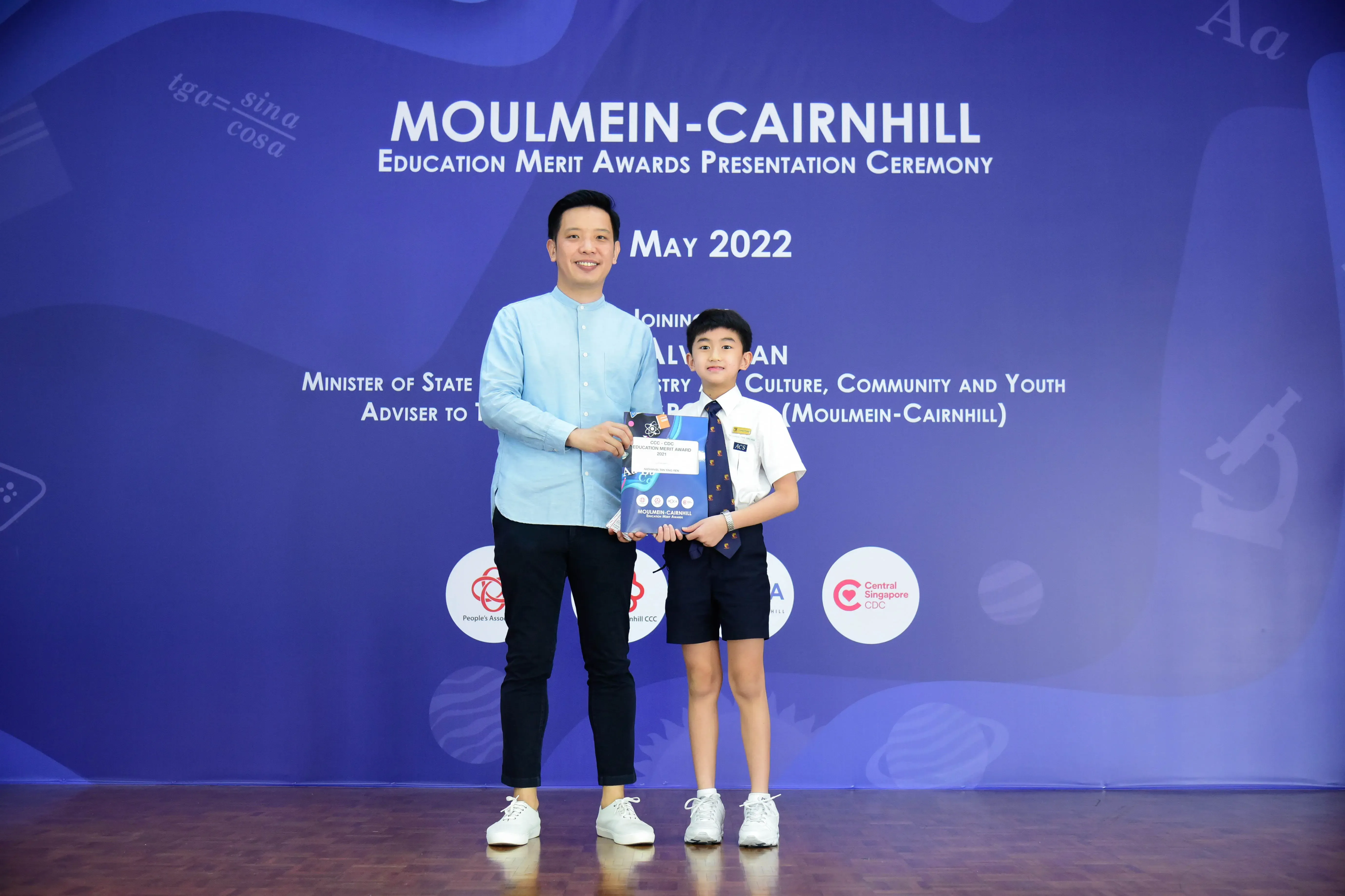 An adult and a young boy in school uniform hold an award together at the Moulmein-Cairnhill Education Merit Awards May 2022.