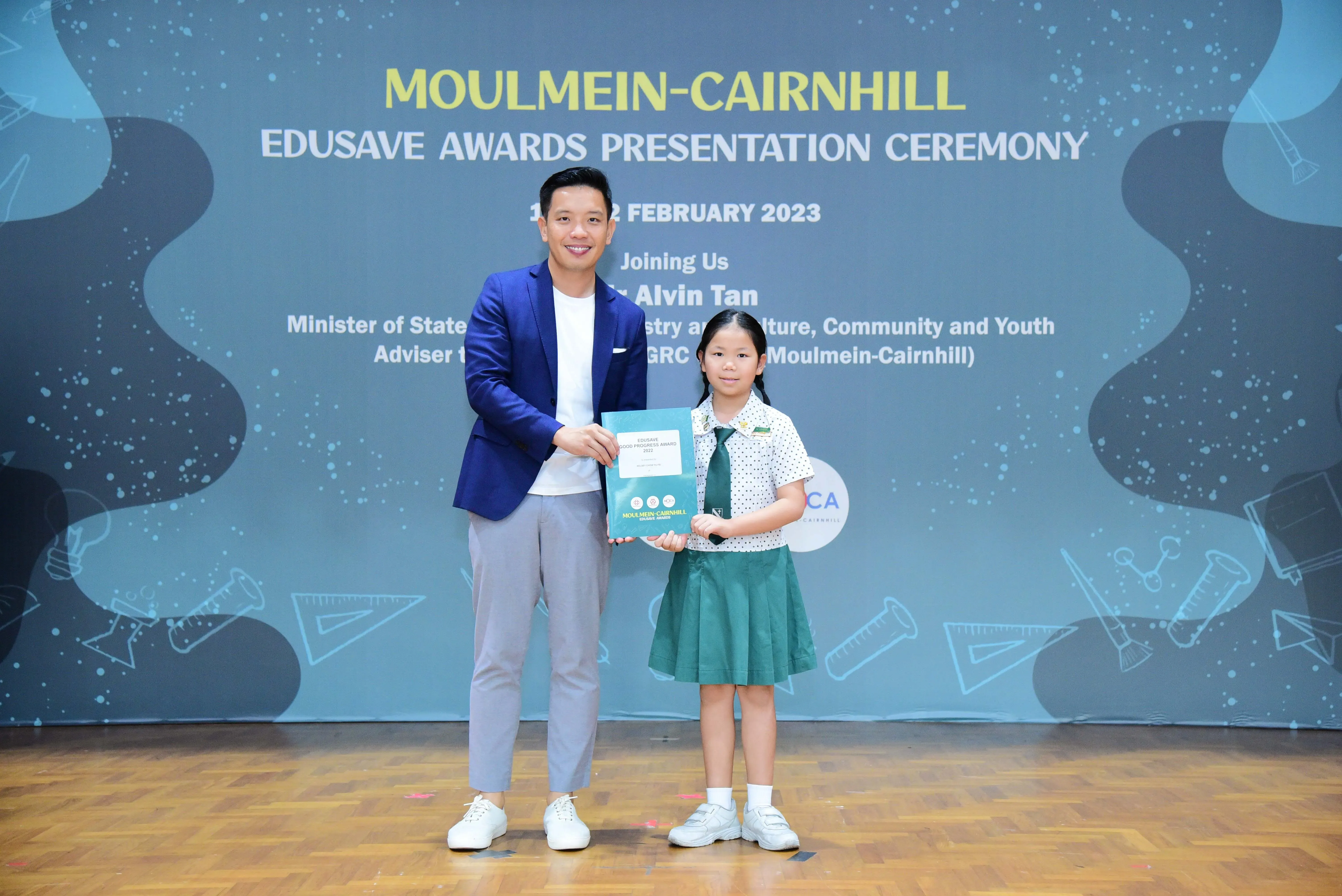 A man in a blue blazer and a young girl in a green school uniform hold an award at the Moulmein-Cairnhill Edusave ceremony.