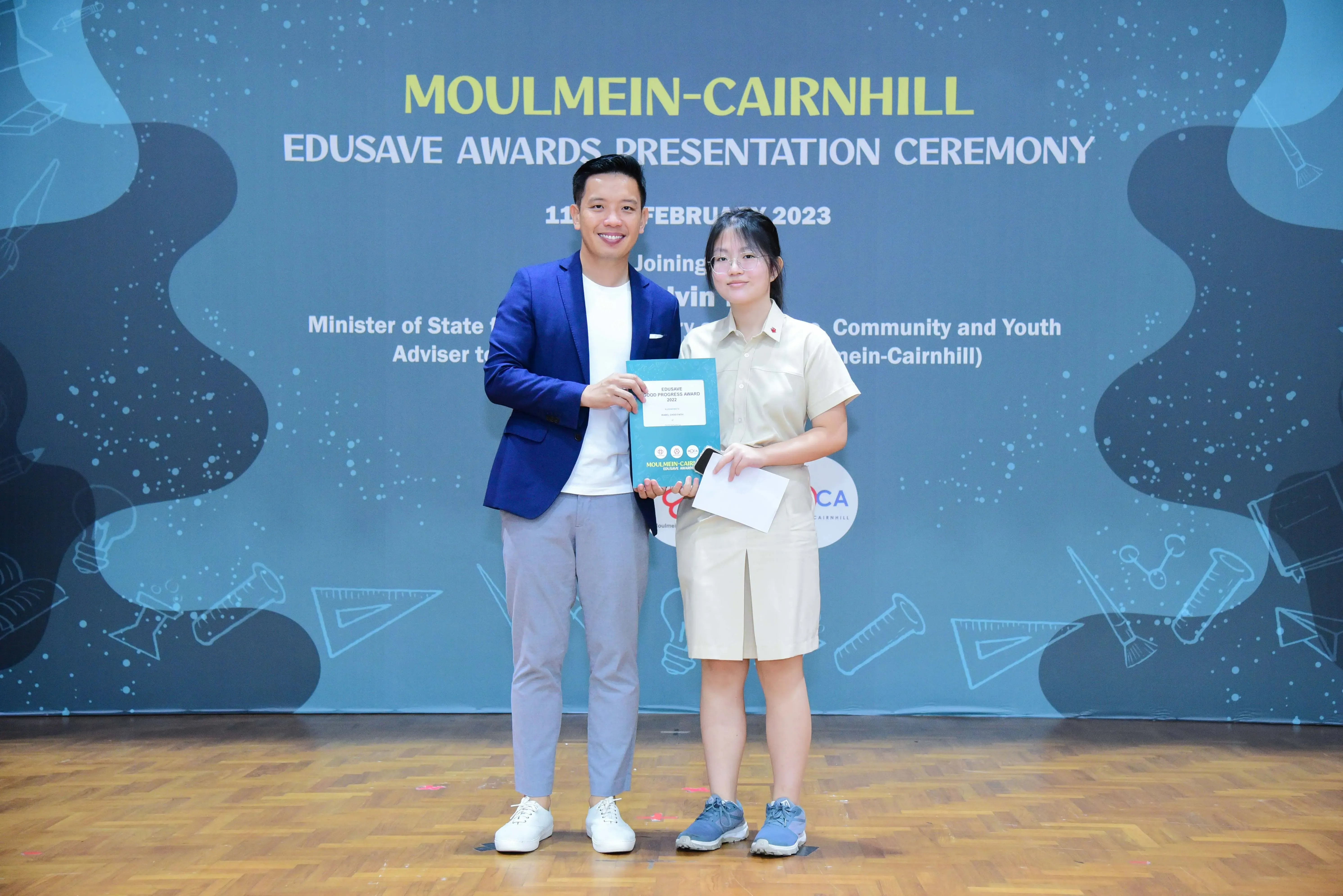 A man in a blue blazer and a student in uniform pose holding an award at the Moulmein-Cairnhill Edusave Awards Ceremony 2023.
