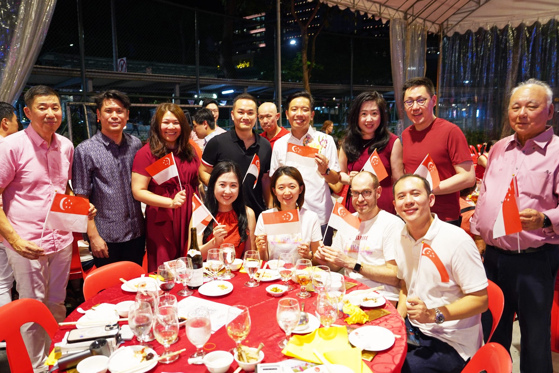 A group of smiling people holding Singapore flags at an outdoor dinner event at night.