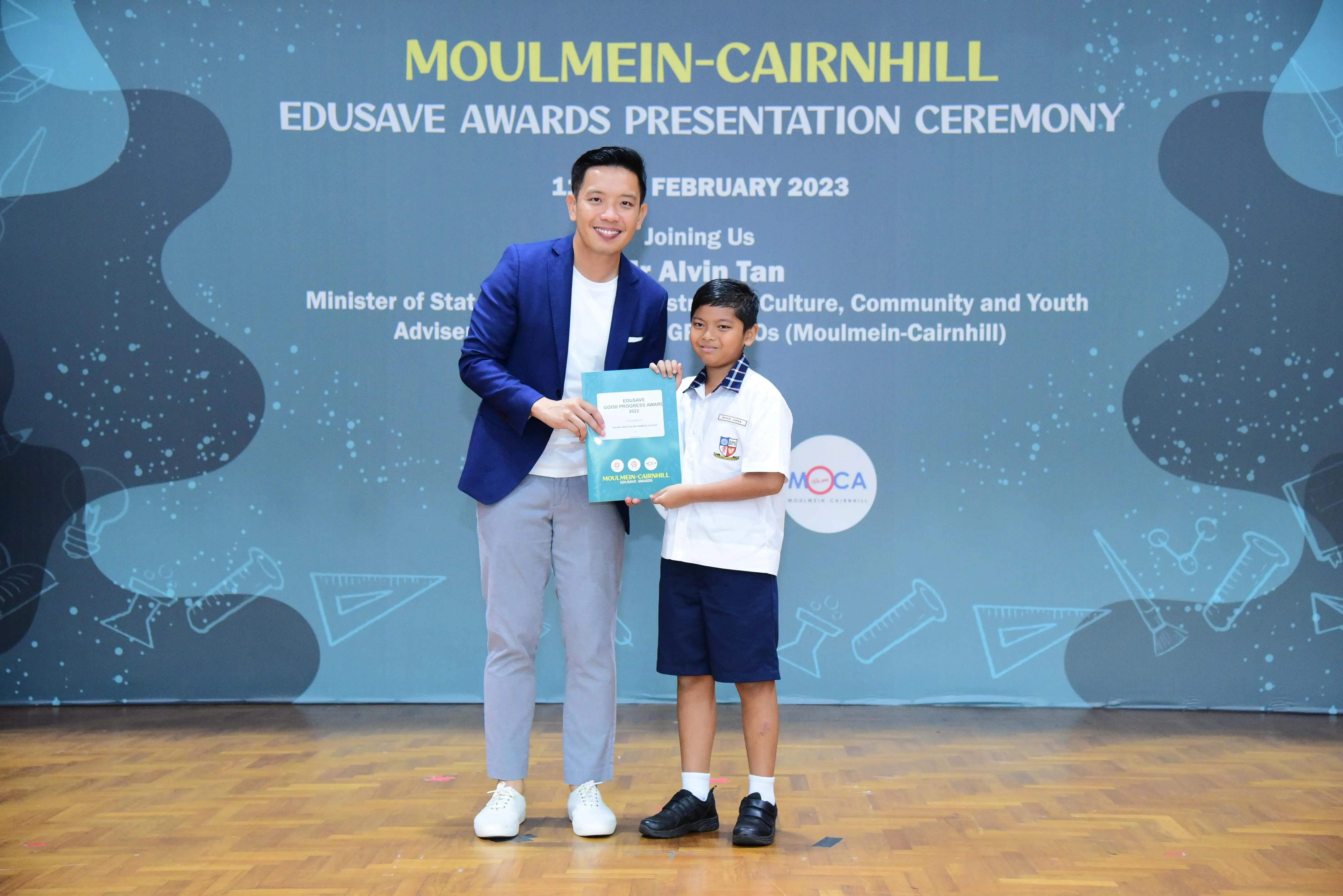 An adult in a blue blazer and a young student in uniform hold a certificate at the Moulmein-Cairnhill Edusave Awards 2023.
