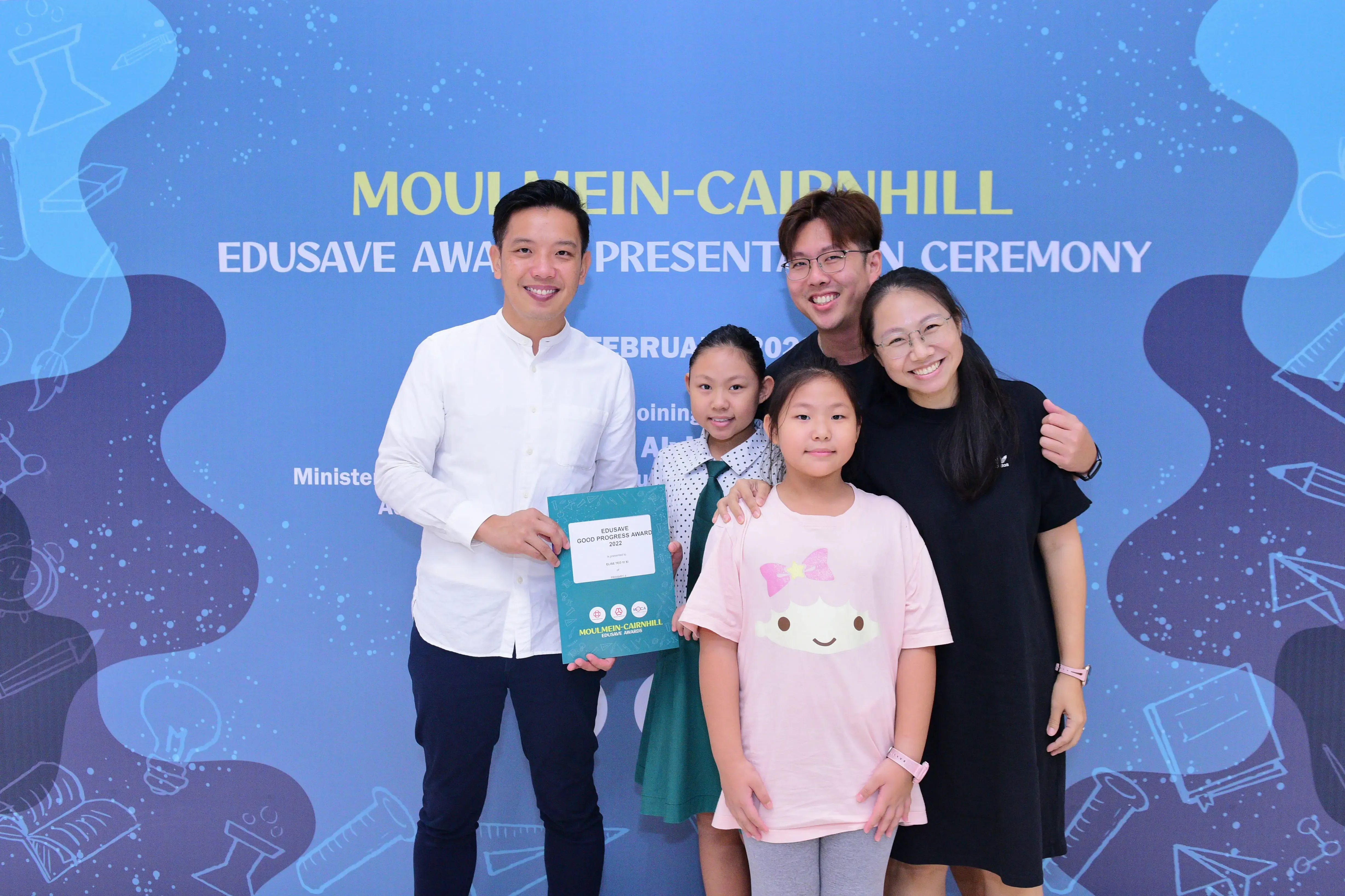 Five smiling people pose at the Moulmein-Cairnhill Edusave Award Presentation Ceremony, holding an award folder.
