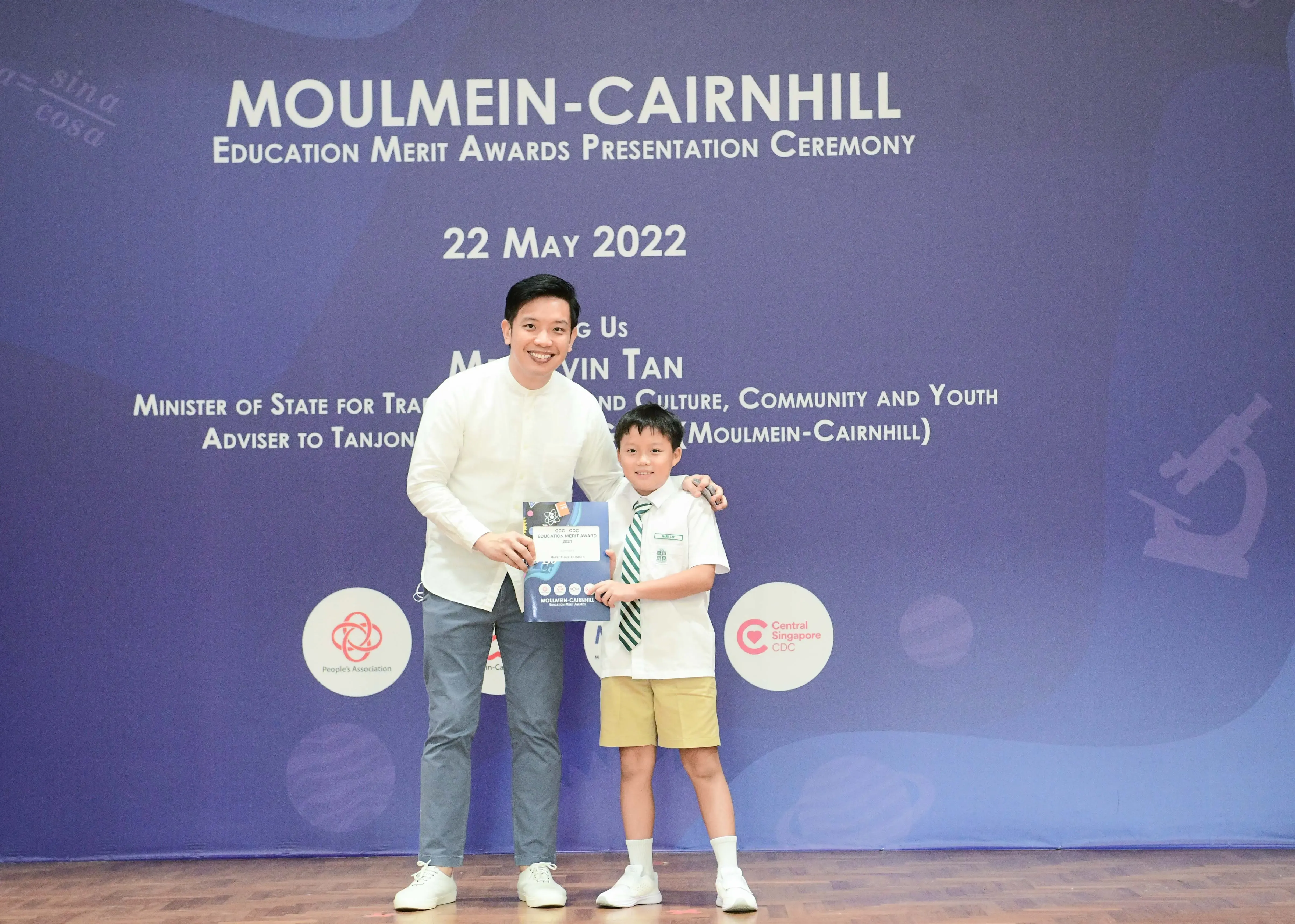 A man and a young boy in school uniform pose together holding an award at the Moulmein-Cairnhill Merit Awards Ceremony.